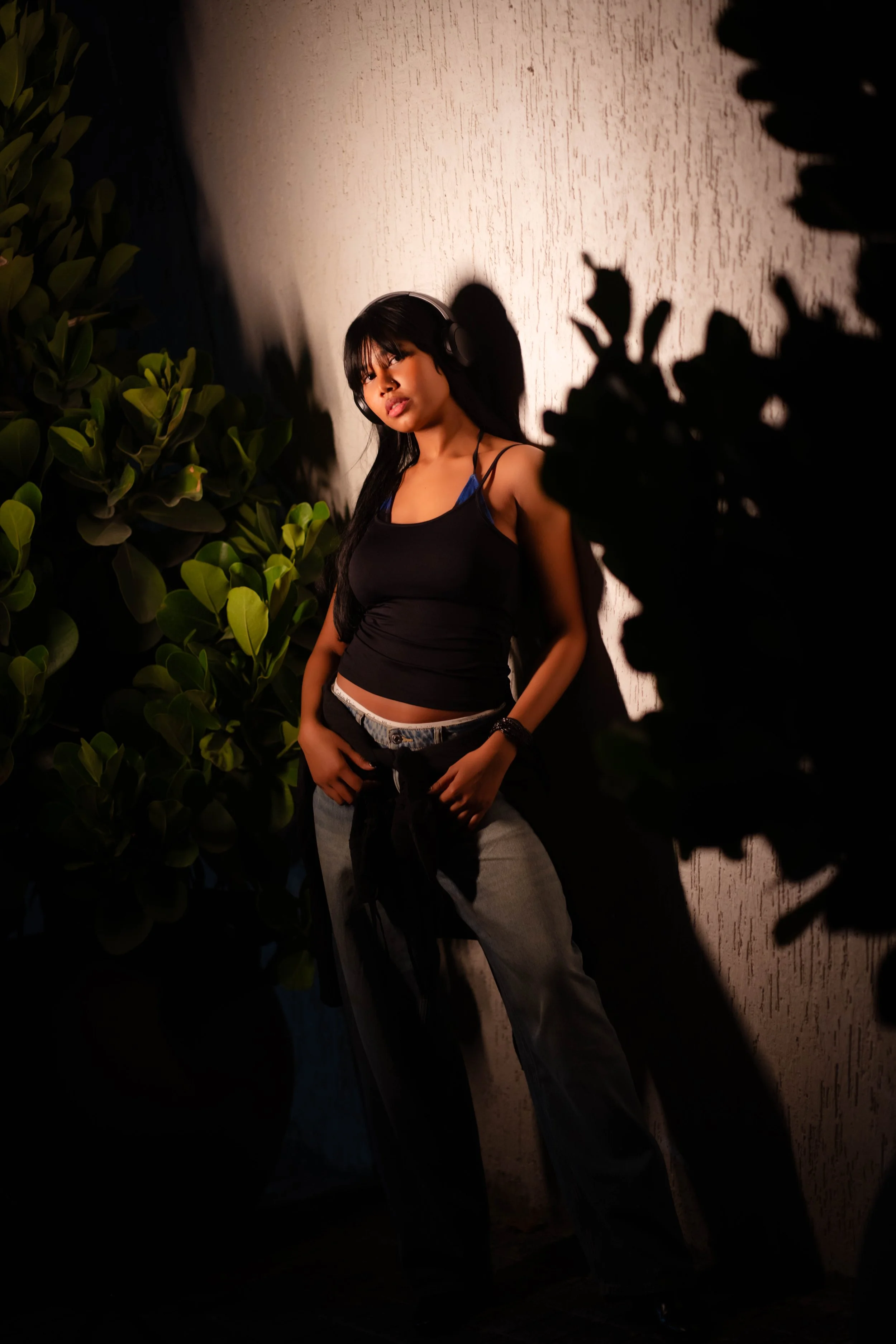 Young woman with long black hair wearing a black tank top, grey sweatpants, and headphones, standing against a textured beige wall with surrounding green foliage.