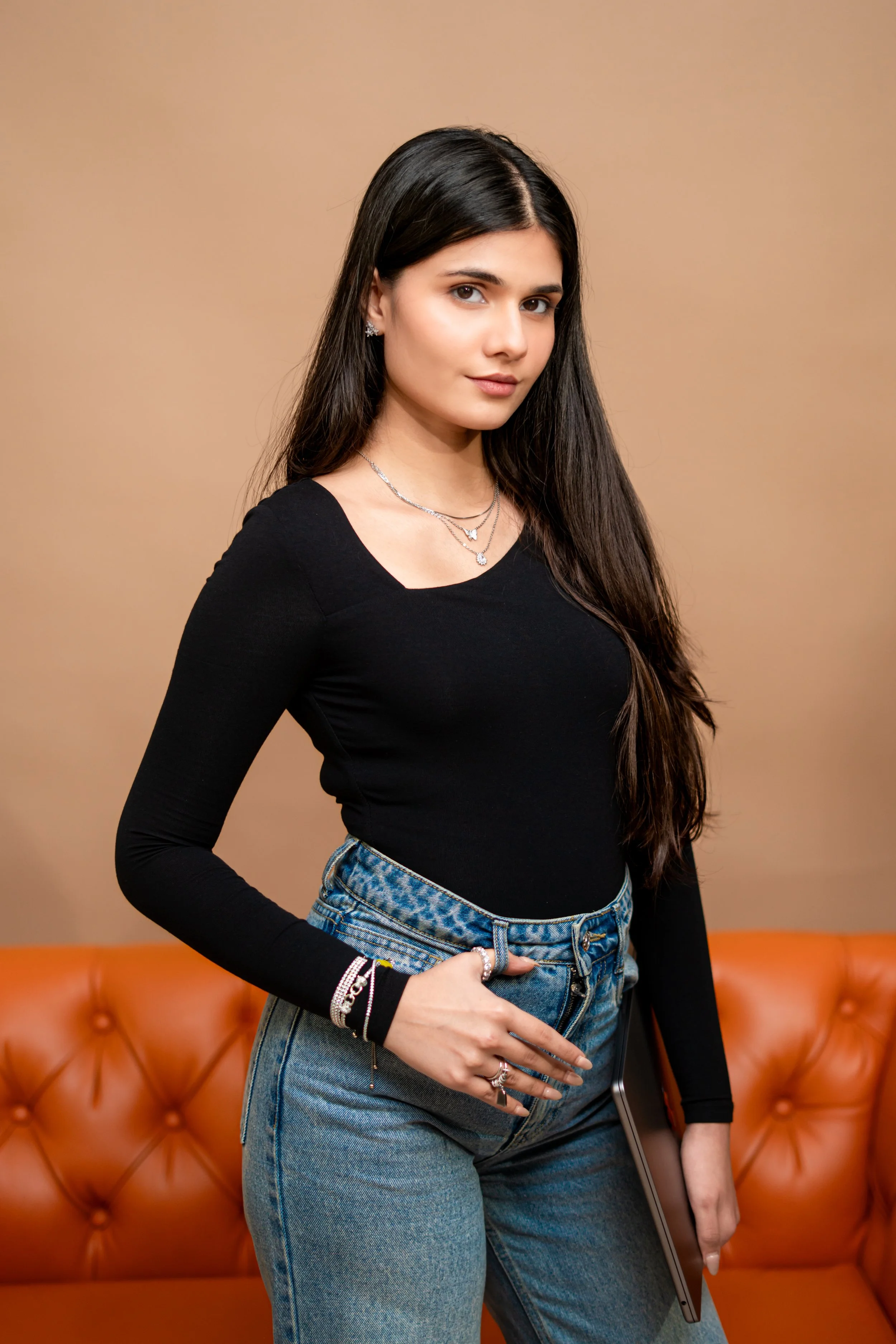 A young woman with long dark hair and fair skin, wearing a black long-sleeve top, light blue high-waisted jeans, and silver jewelry, standing against a beige background with an orange couch in the background.