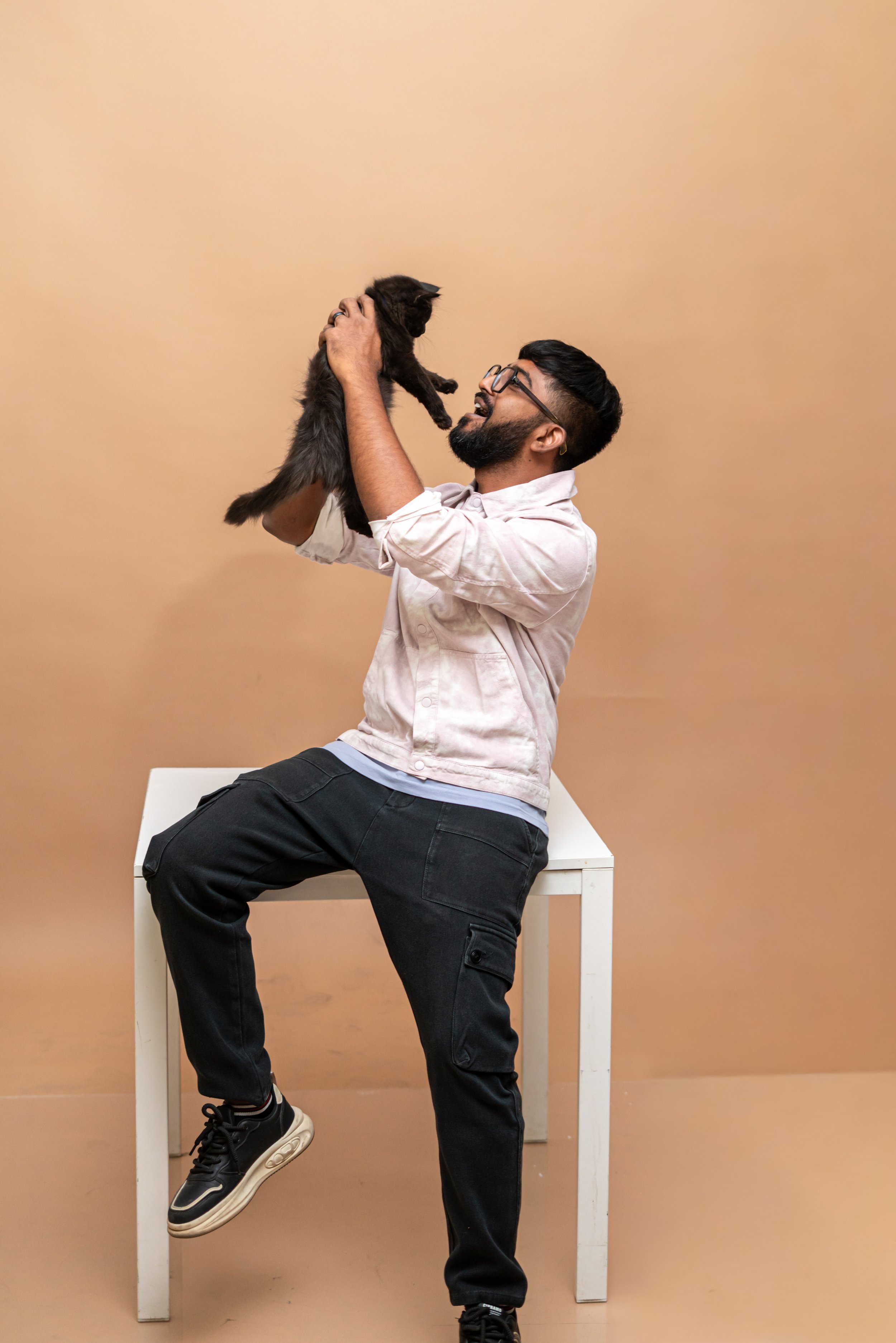 A man with glasses, beard, and dark hair, wearing a light pink jacket, black pants, and sneakers, sitting on a white table, holding a black kitten up in the air, with a beige background.