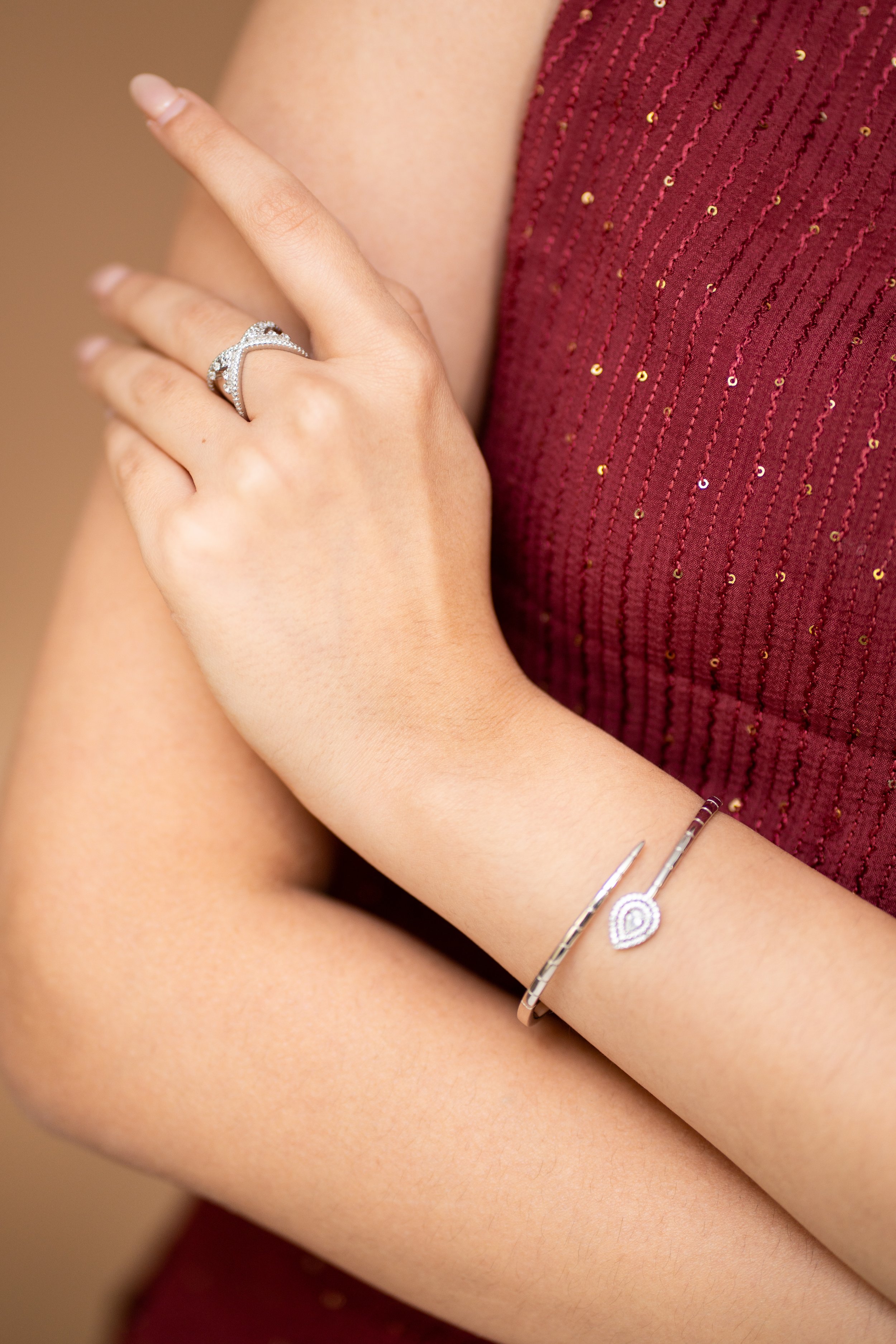 Close-up of a woman's arm with jewelry, including a diamond ring on her finger and a silver bracelet with a teardrop-shaped charm, against a red, sequined dress.