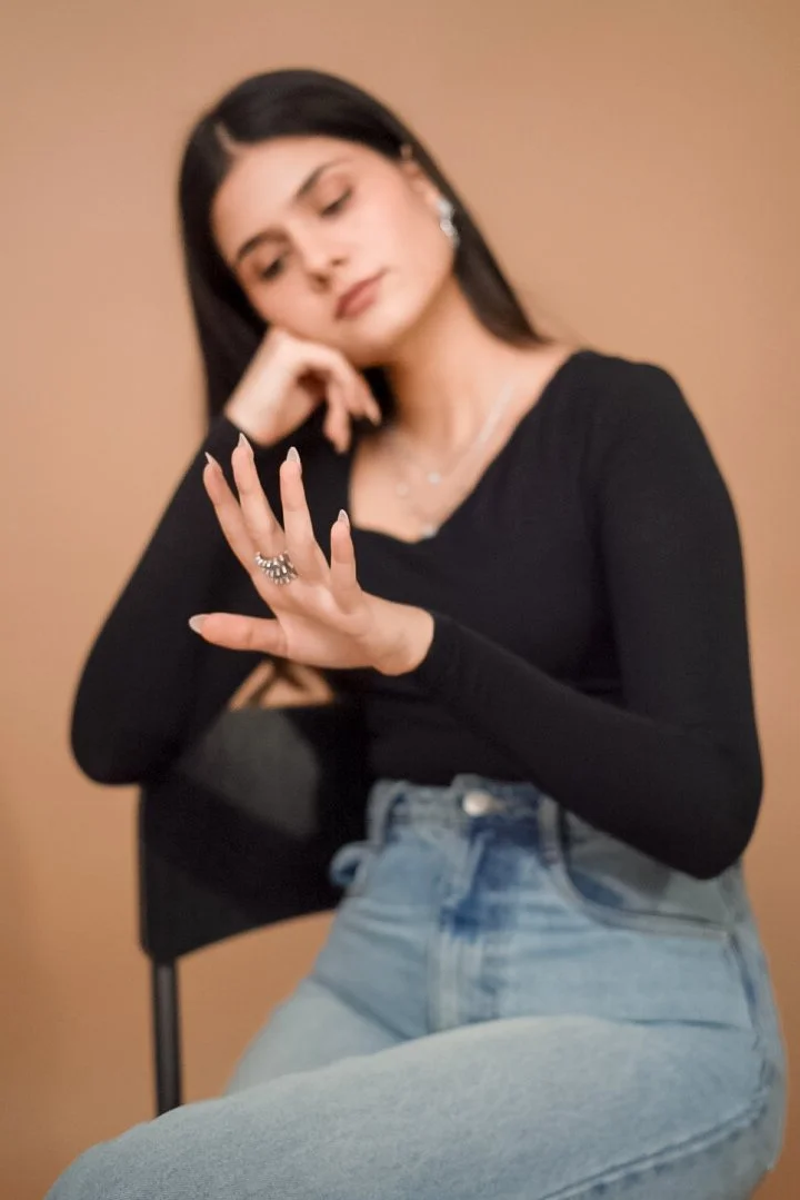 A young woman with long black hair, dressed in a black long sleeve top and light blue jeans, sitting against a plain beige background. She is looking down and touching her forehead with one hand, while her other hand is raised with fingers extended, 