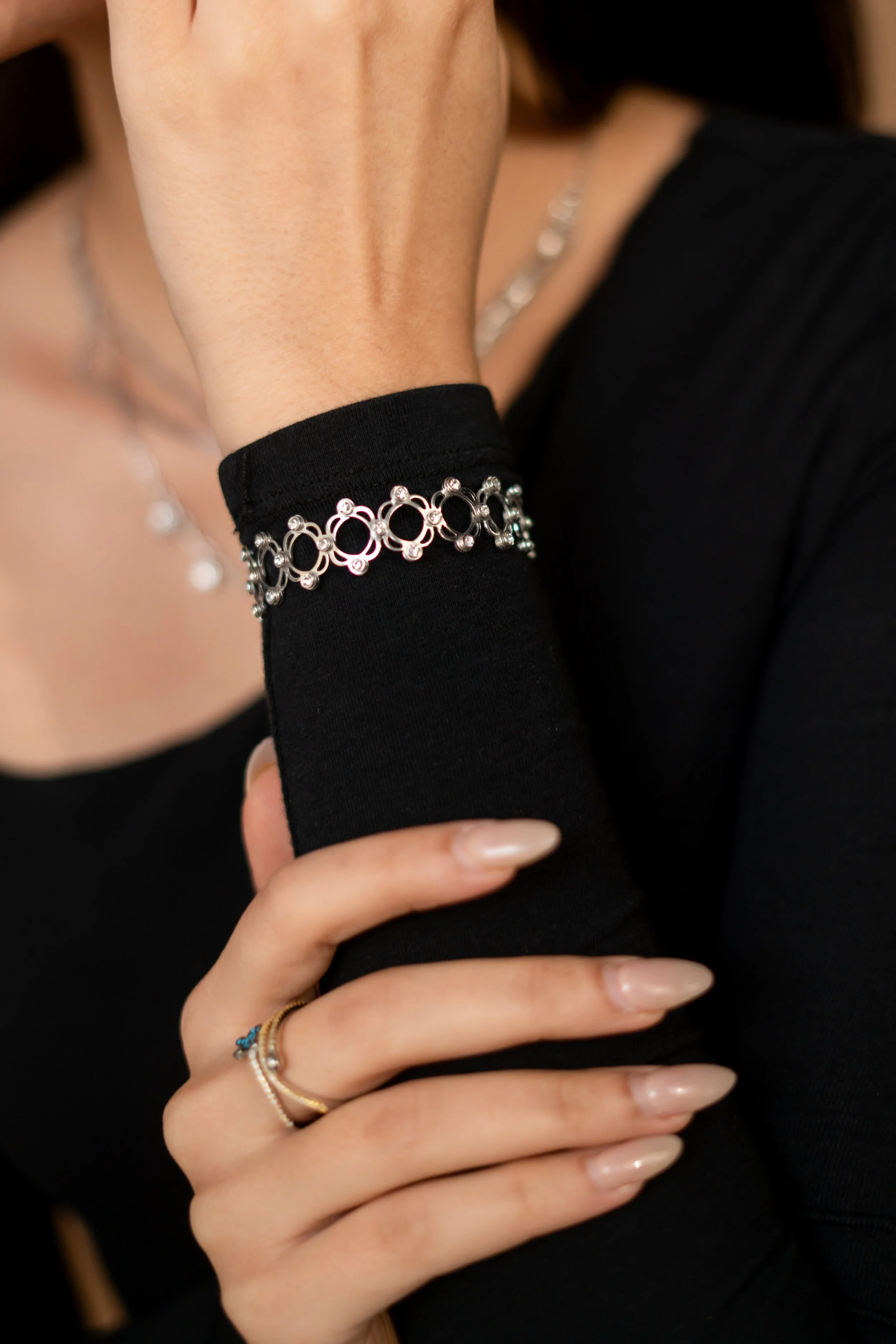 Close-up of a woman's hand and wrist, showing a black long-sleeve shirt, silver jewelry including rings and a bracelet, and her face partly visible in the background.