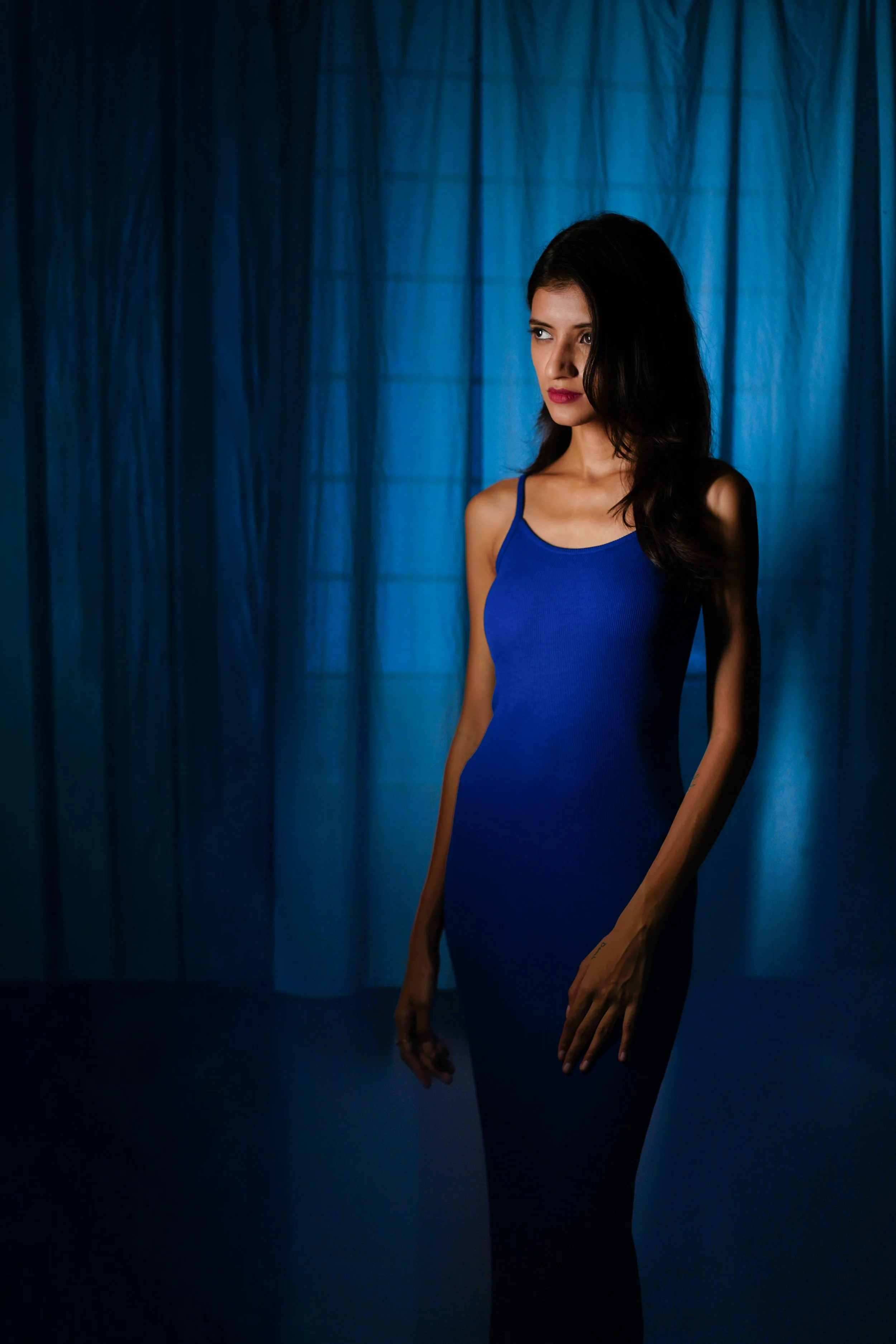 A woman with long dark hair wearing a blue sleeveless dress standing in front of a blue curtain background.