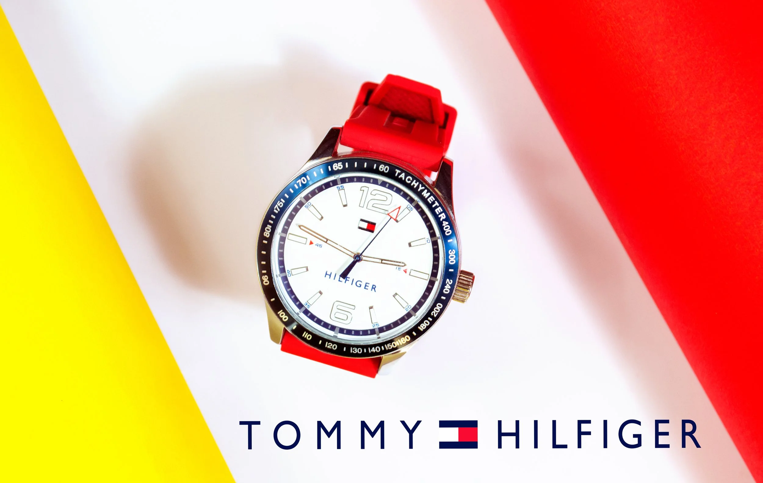 A Tommy Hilfiger wristwatch with a white face, black bezel, and red strap, placed on a background composed of yellow, white, and red sections, with the Tommy Hilfiger logo at the bottom.