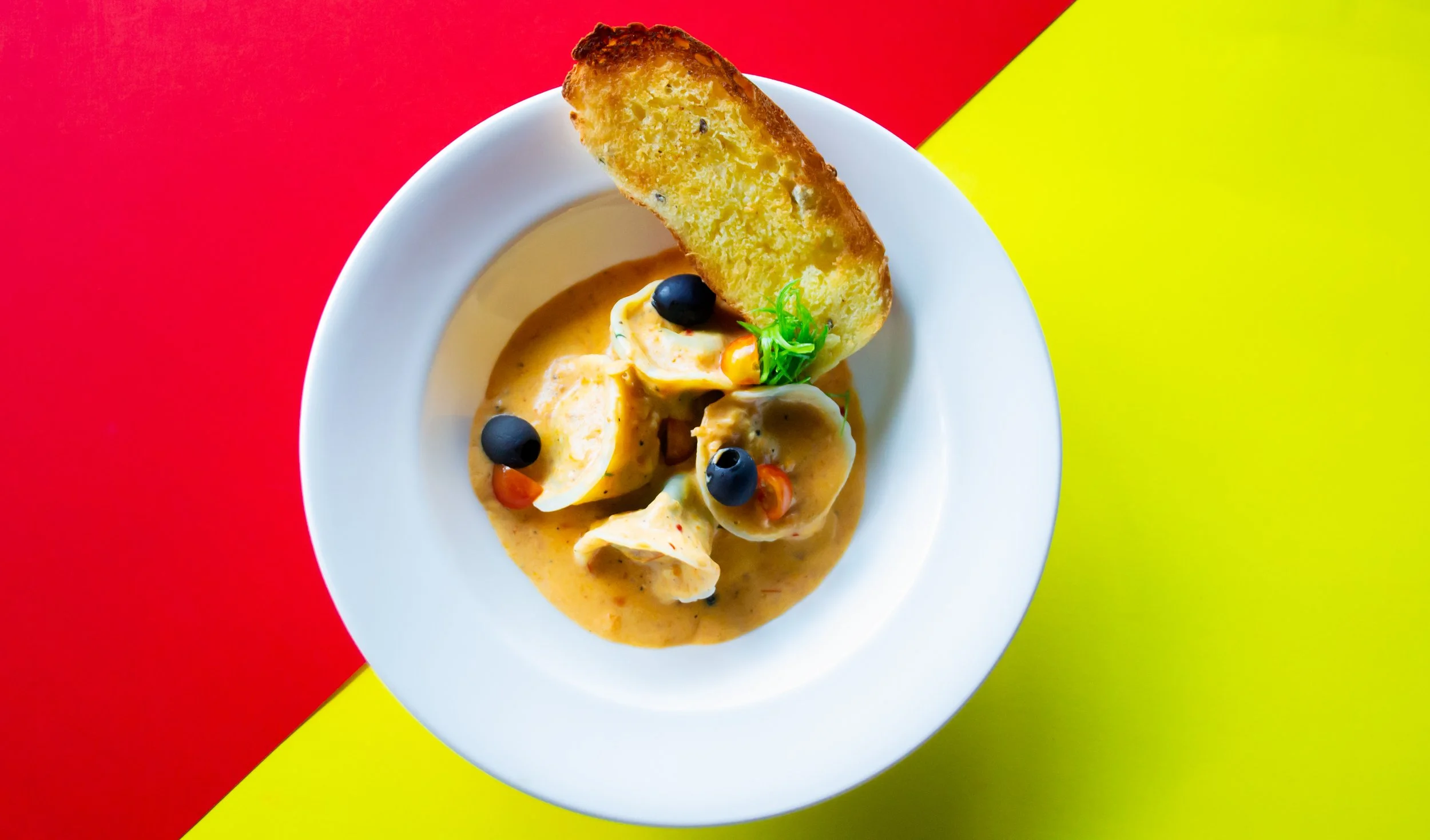 A plate of eggs in a creamy sauce garnished with black olives, red peppers, and a sprig of green herbs, served with a slice of toasted bread on the side, on a background split diagonally between red and yellow.