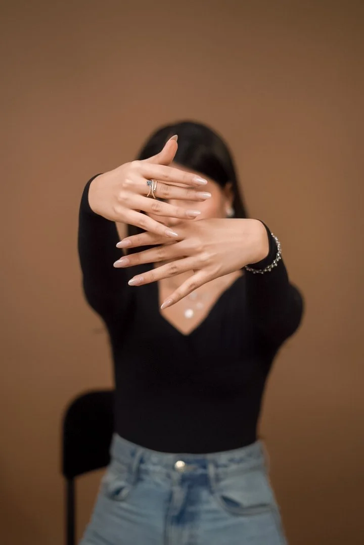 A woman with dark hair wearing a black top and jeans, covering her face with her hands, showcasing rings and bracelets against a brown background.