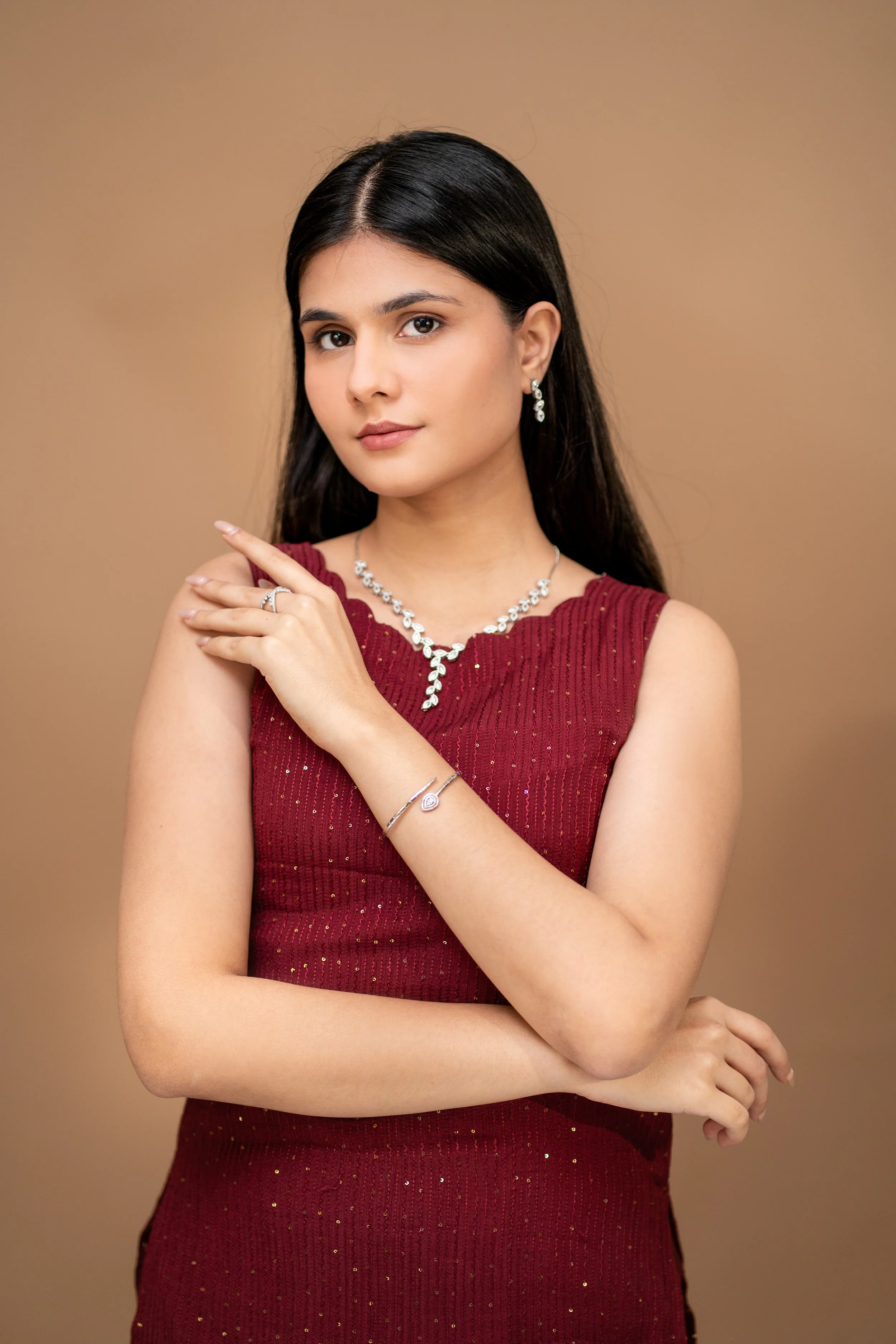 A woman with long dark hair wearing a sleeveless red dress with gold accents, pearl earrings, and a matching necklace, posing with her right hand on her left shoulder against a neutral background.