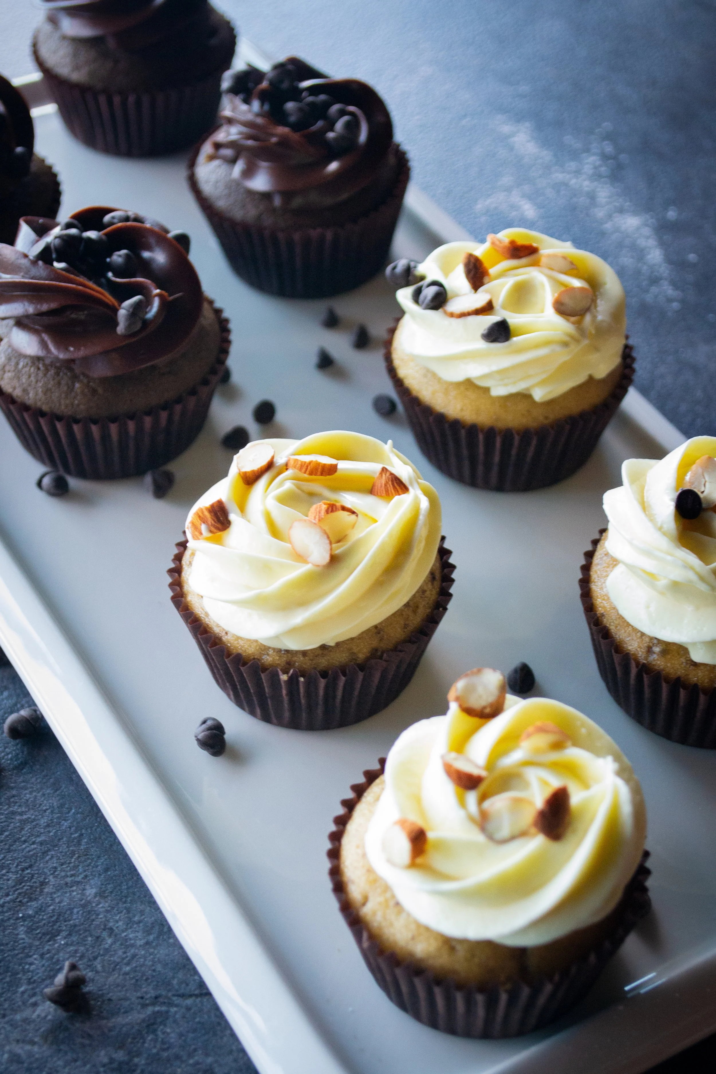Assorted cupcakes with chocolate frosting and white frosting topped with nuts and chocolate chips on a white platter.