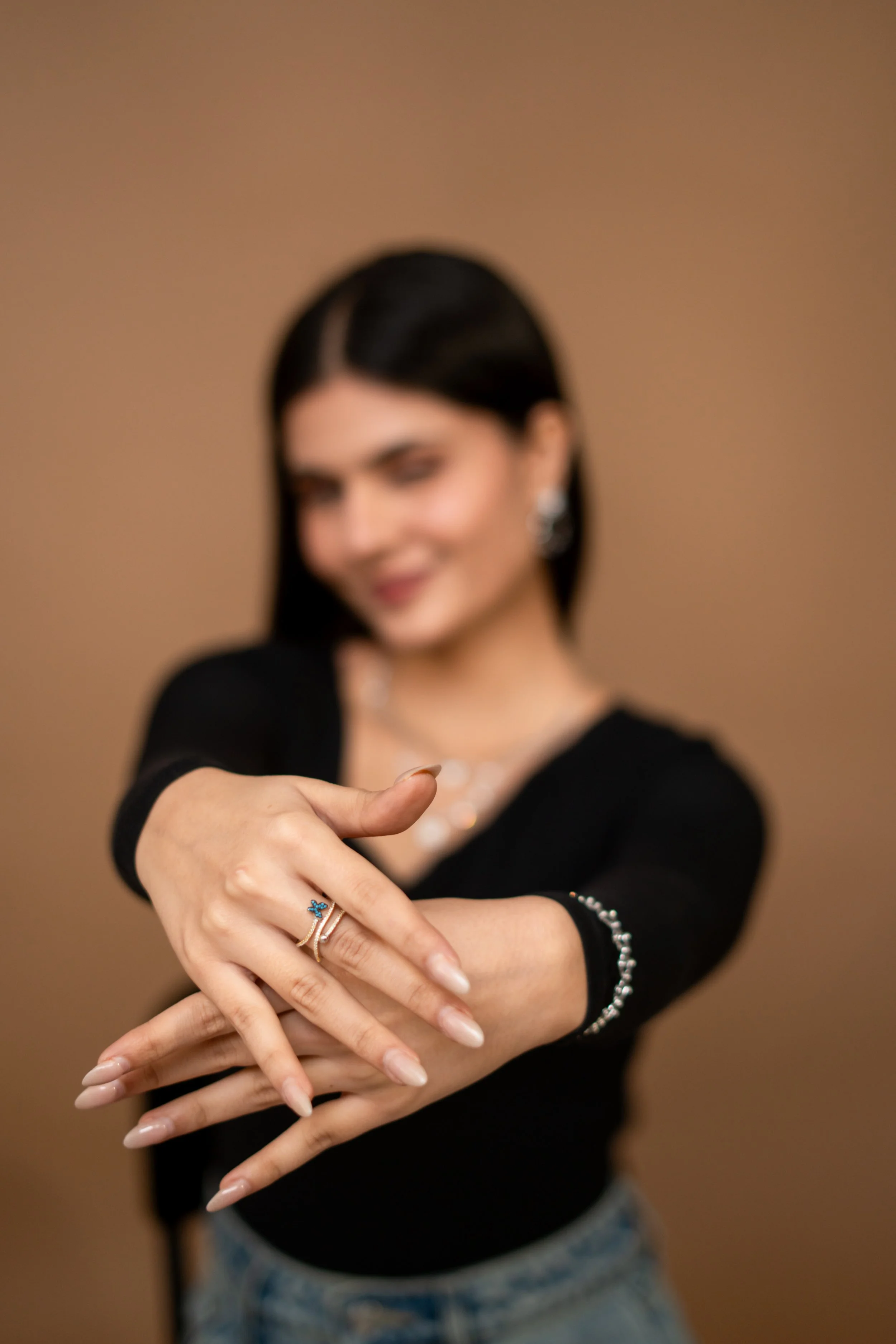 A woman with dark hair wearing a black top, showing her hands with jewelry, including rings and a bracelet, against a beige background.