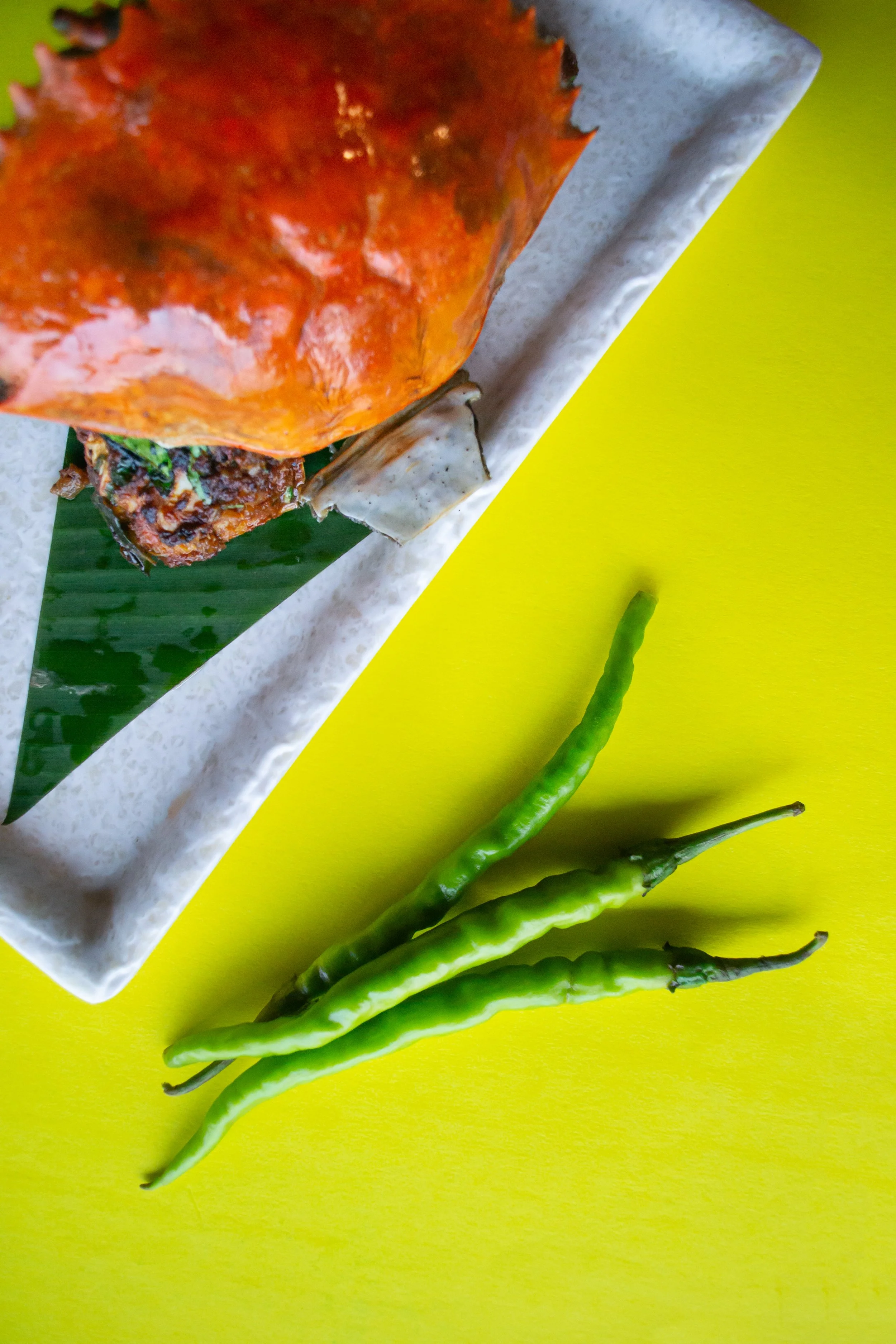 Three green chili peppers beside a white rectangular dish with a partially visible spicy, saucy dish on a yellow surface.
