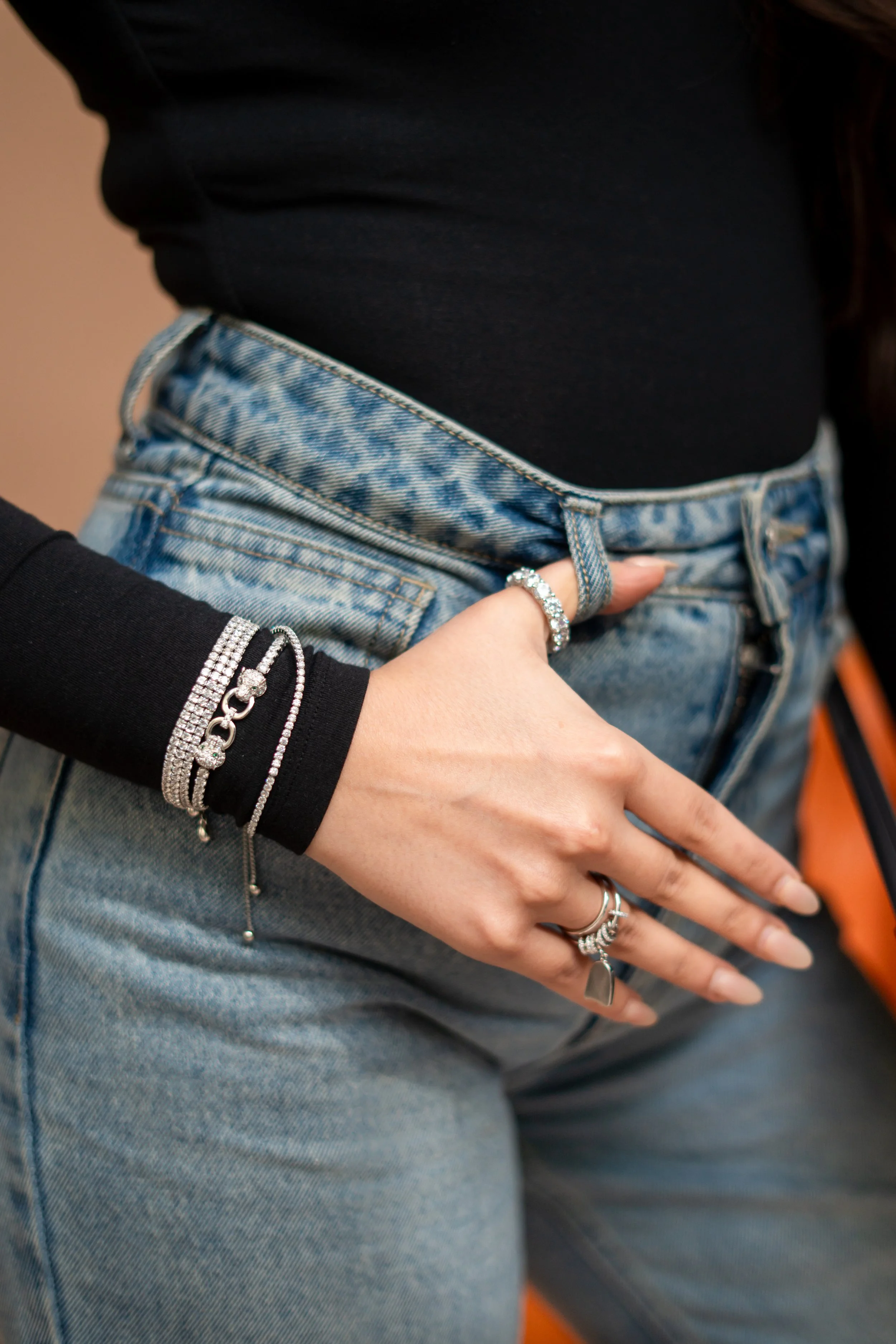 Close-up of a woman's midsection showing her wearing a black top and light blue high-waisted jeans. She has multiple rings and bracelets on her left hand, which is resting near her waist.