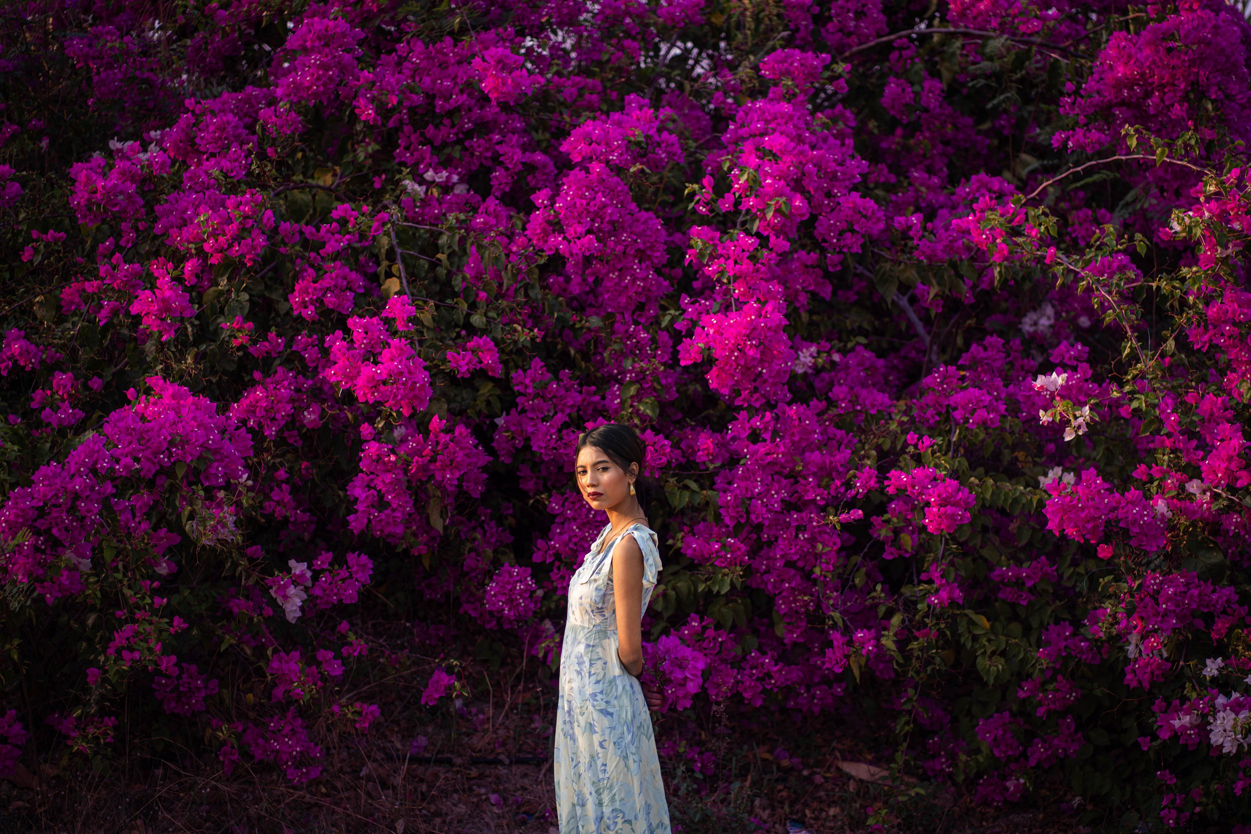 A woman in a light-colored floral dress stands in front of a large, vibrant bush of pink bougainvillea flowers.