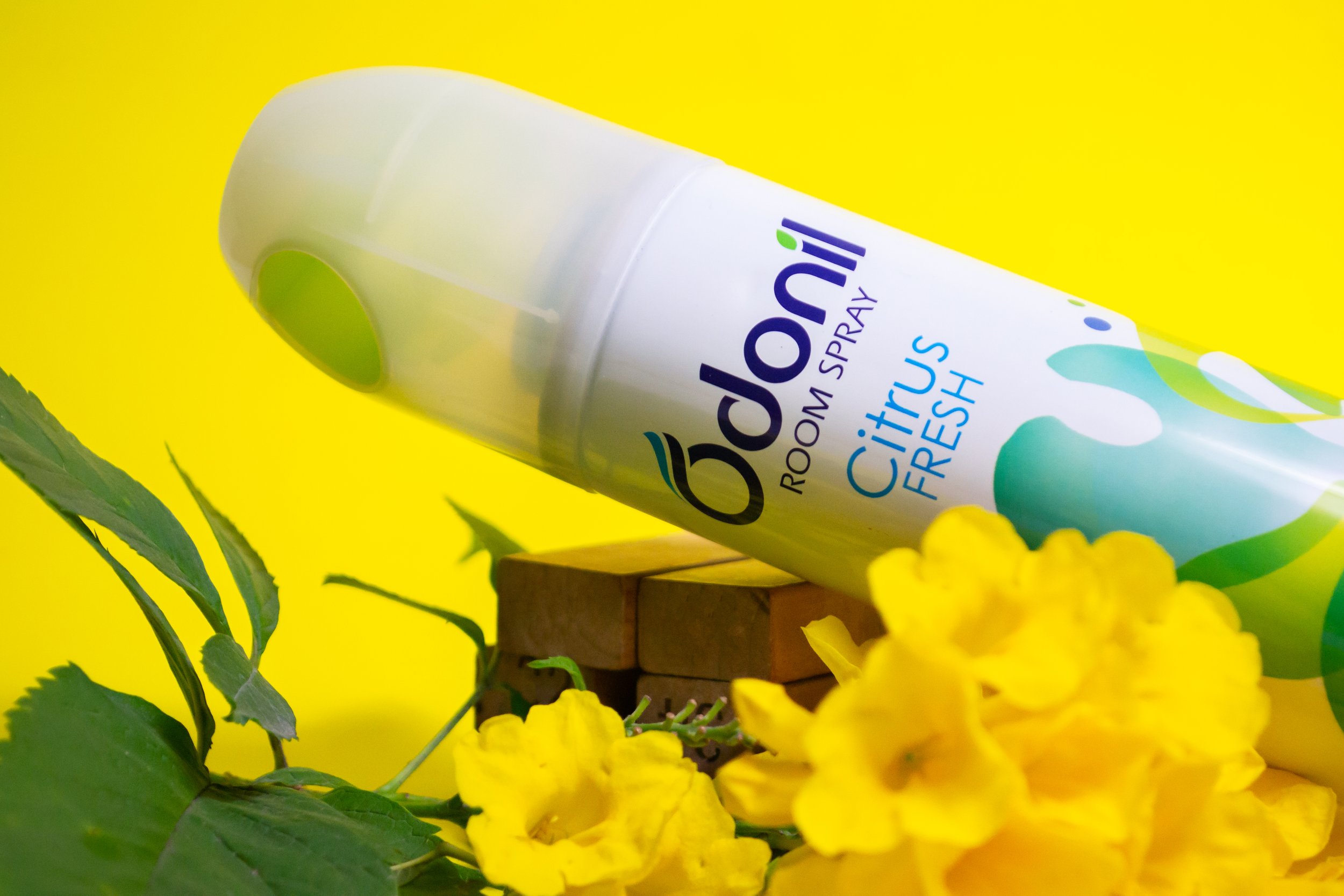 A Bedlam Room Spray can with Citrus Fresh scent resting on a small wooden block, accompanied by yellow flowers and green foliage against a bright yellow background.