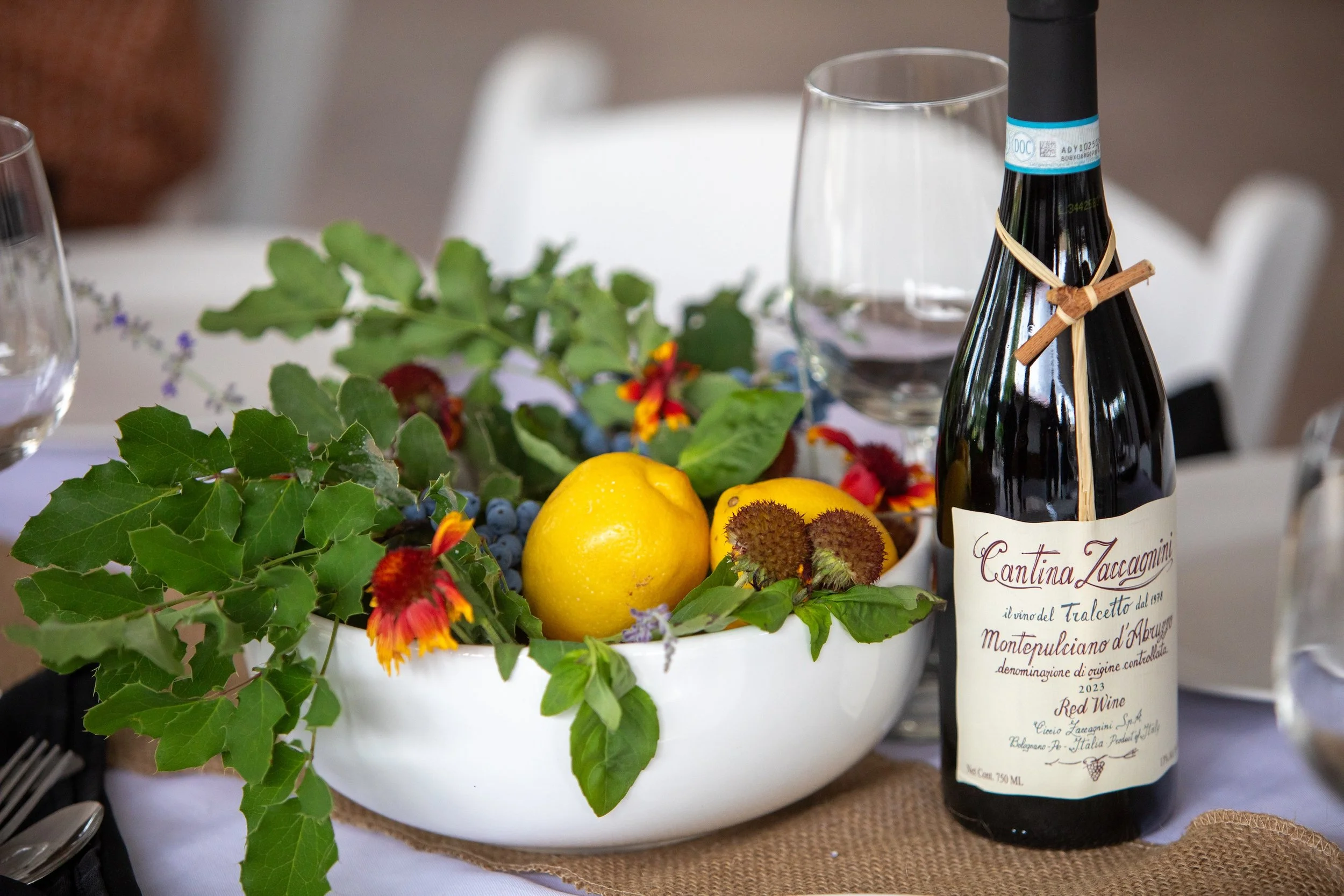 A wine bottle labeled 'Cantina Zaccagnini' placed next to a white bowl filled with green leaves, yellow lemons, and some flowers. Two empty glasses are behind the bowl, on a table set for a meal.