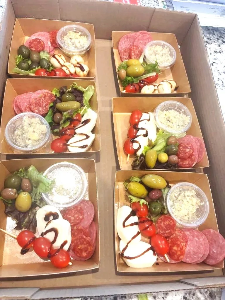 Six small cardboard trays filled with assorted charcuterie, cheeses, cherry tomatoes, green and black olives, and a small container of dipping sauce.
