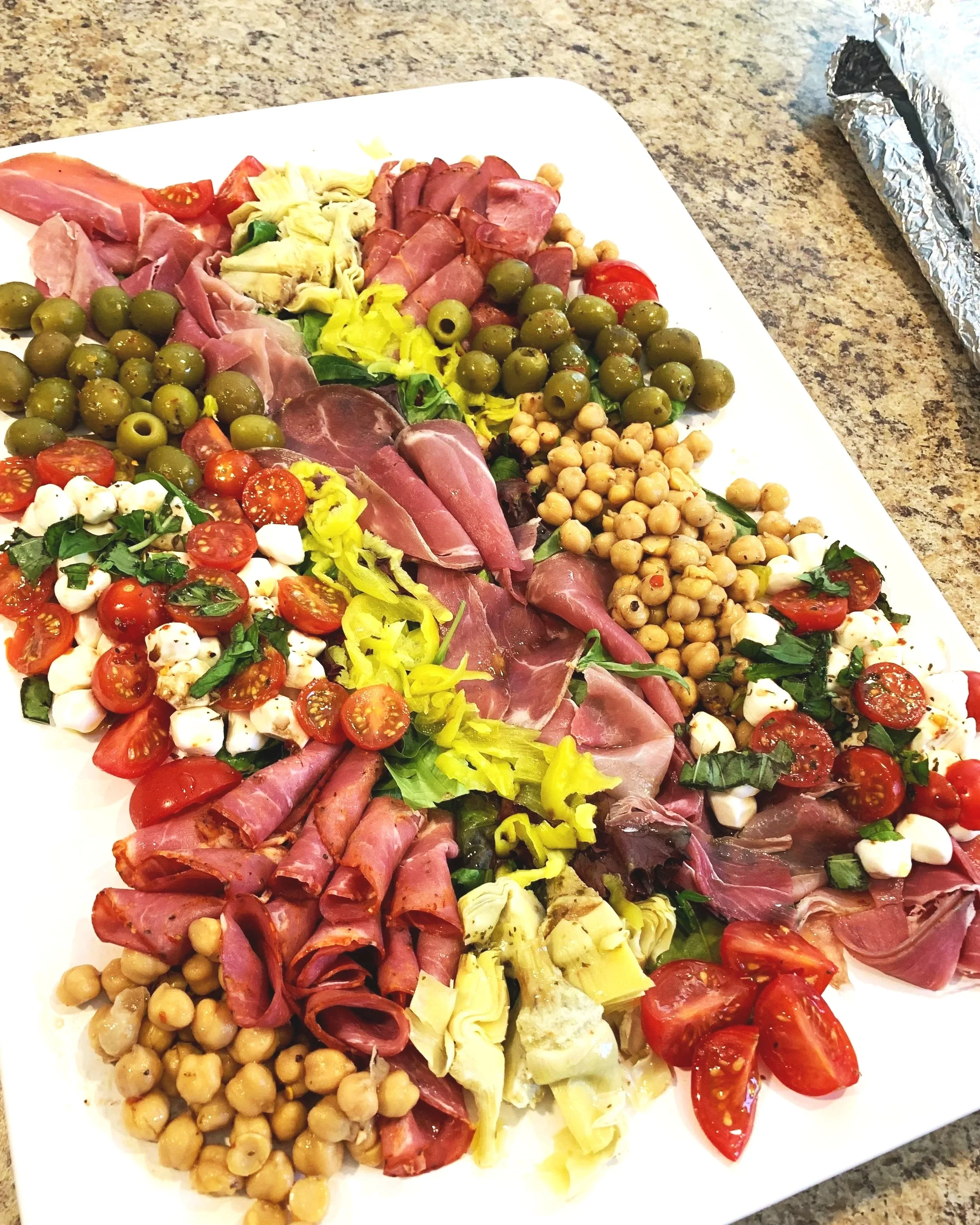 A white rectangular platter filled with assorted charcuterie and vegetable toppings, including cherry tomatoes, mozzarella balls, chickpeas, olives, various cured meats, and leafy greens, set on a granite countertop.