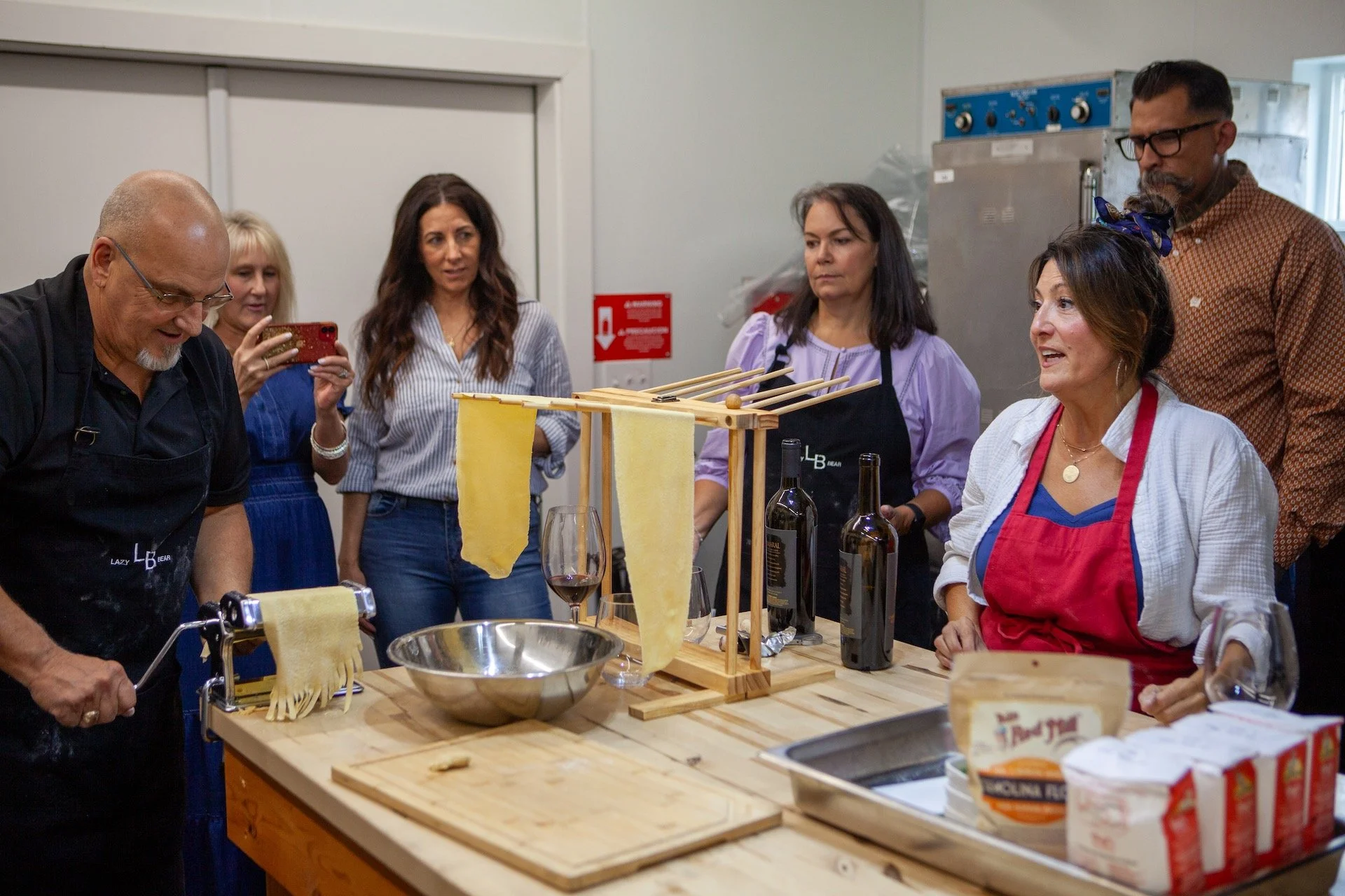 Group of people gathered around a wooden table in a kitchen, participating in a pasta-making class. Some are observing, one person is taking a photo, and others are smiling and talking. Bottles of wine, a metal bowl, and pasta-making equipment are on the table.