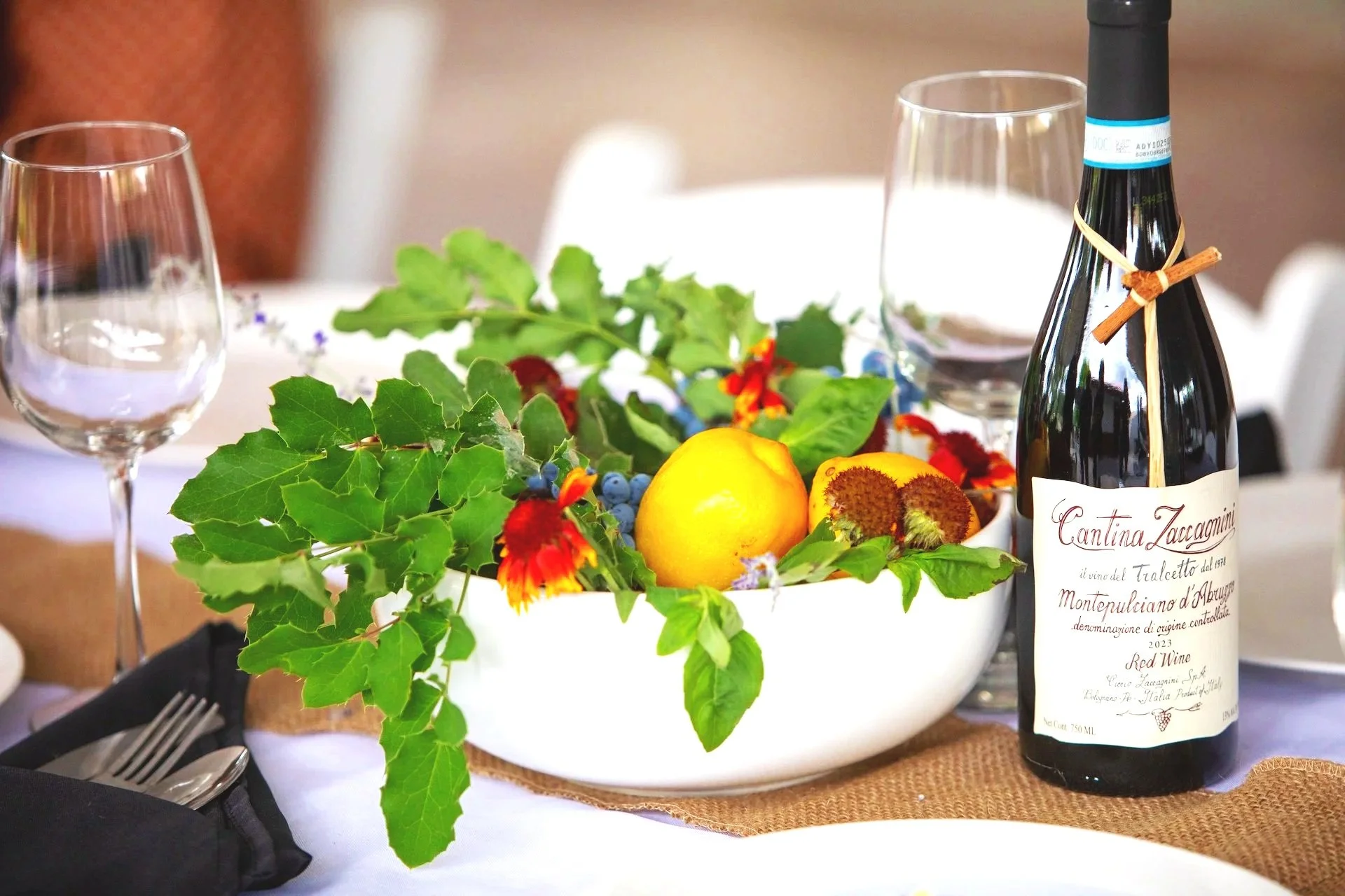 A table decorated with a white bowl of fruit including lemons and peaches, surrounded by colorful flowers and green leaves, with a bottle of red wine labeled 'Cantina Zaccagnini' and a glass of red wine, set with wine glasses, cutlery, and black napkins.