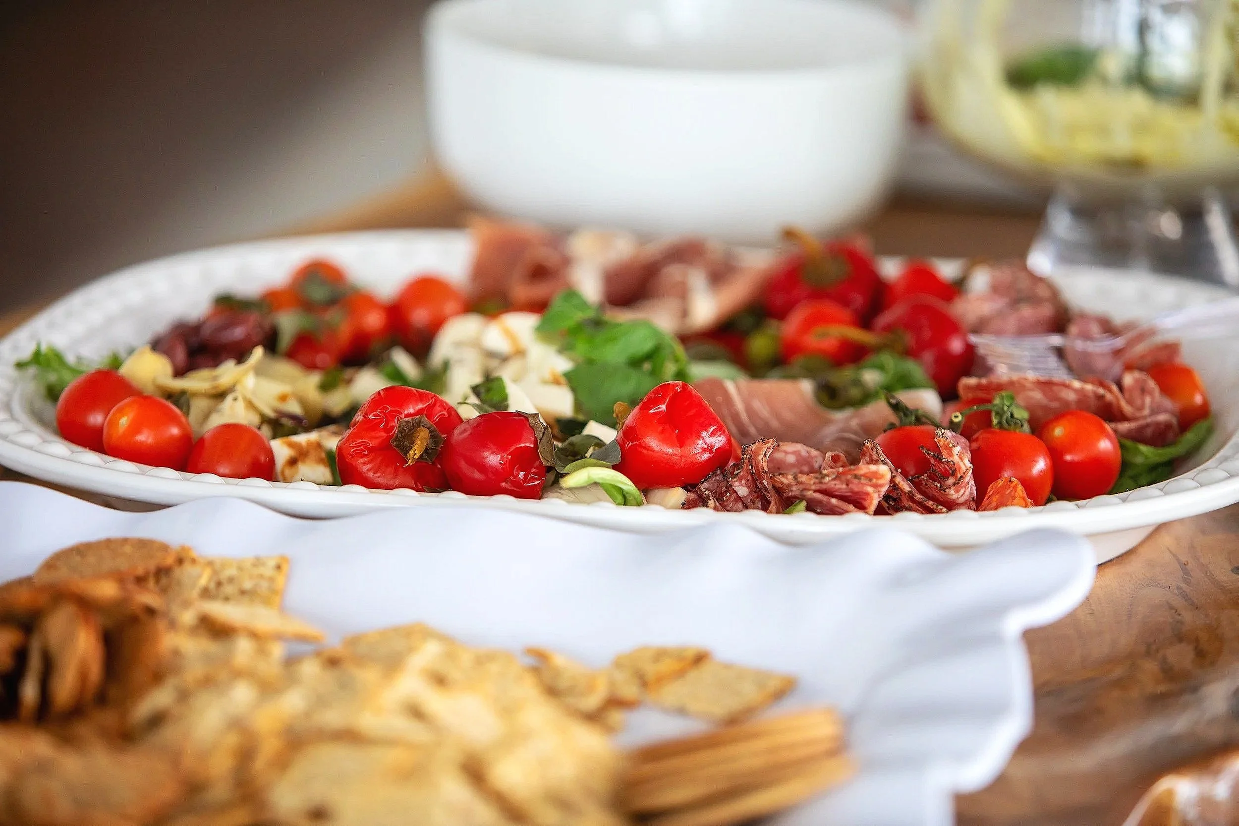 A white serving platter filled with cherry tomatoes, sliced cheese, herbs, and cured meats, surrounded by other dishes in the background.