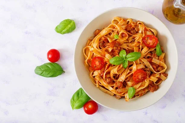 A bowl of cooked pasta with cherry tomatoes, basil leaves, and ground meat, garnished with fresh basil, on a white surface.