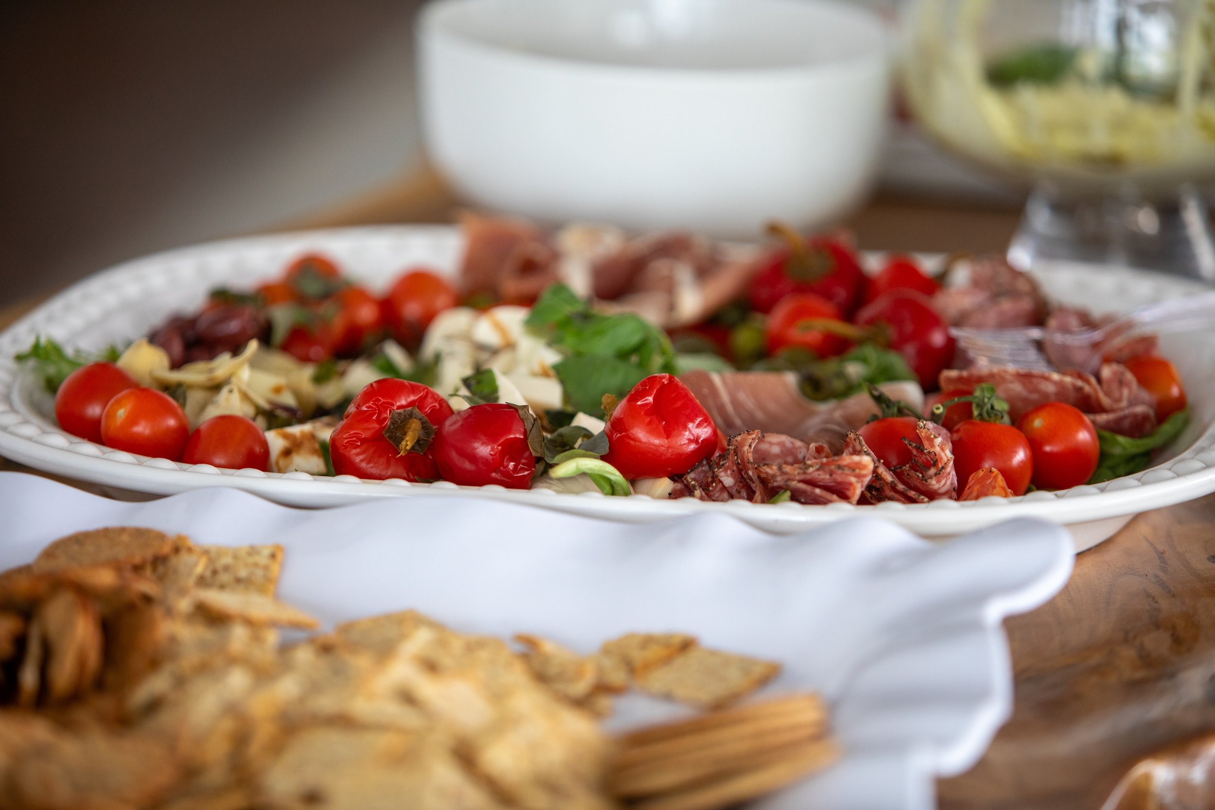 A white platter filled with cherry tomatoes, basil, cheese, and cured meats, with other dishes and bowls visible in the background on a wooden table.