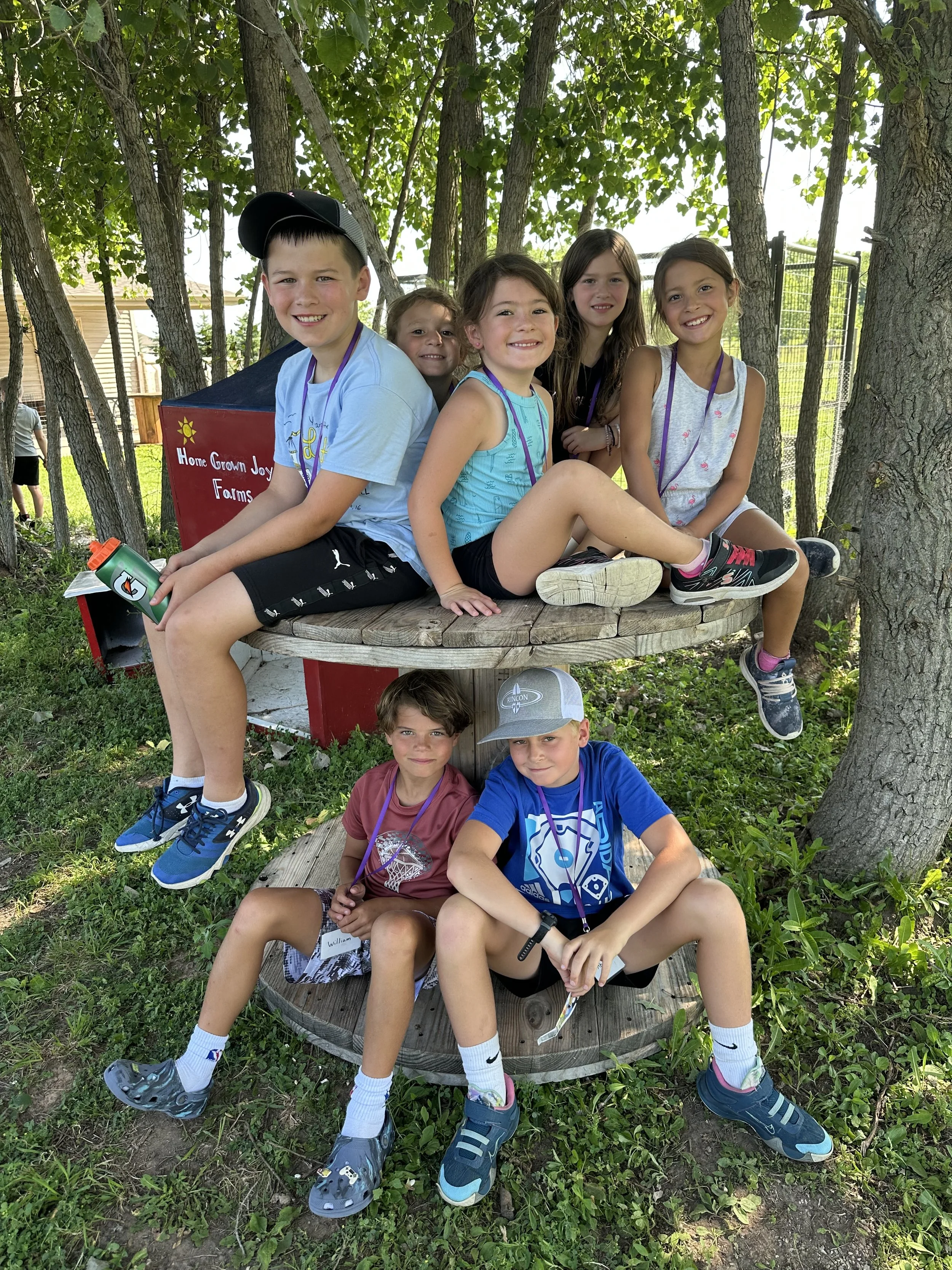 Group of seven children sitting and standing on and around a round wooden platform outdoors in a wooded area with trees and a fence, smiling at the camera.
