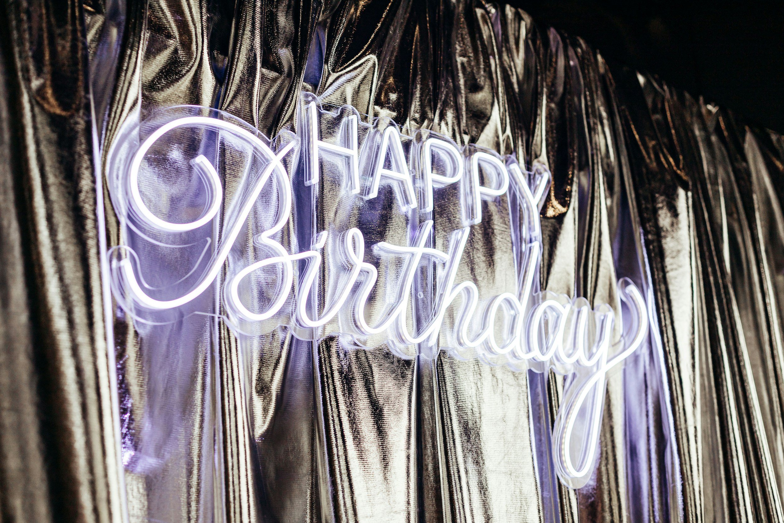 Neon sign reading 'Happy Birthday' in front of a metallic curtain.