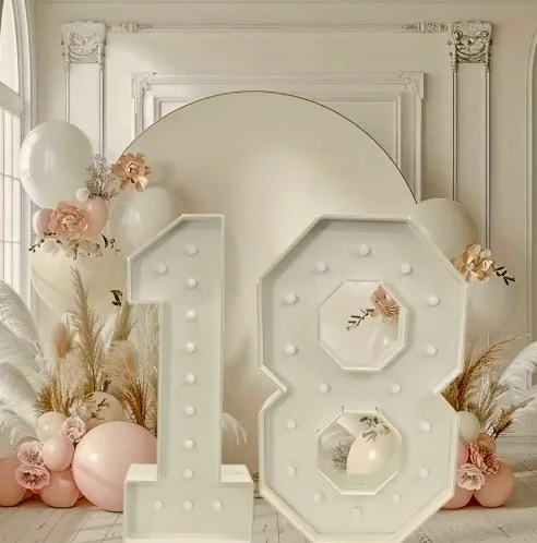 Large white illuminated number 18 decorated with pink and white balloons, flowers, and pampas grass for a celebration or birthday party in a decorated room.