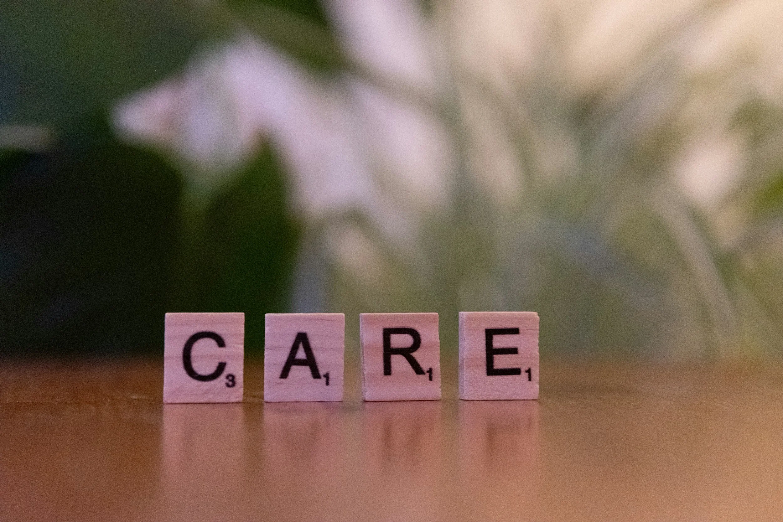 Four wooden Scrabble tiles spelling out the word "CARE" on a wooden surface with a soft-focus floral background. Delta Doc, DPC is about CARE