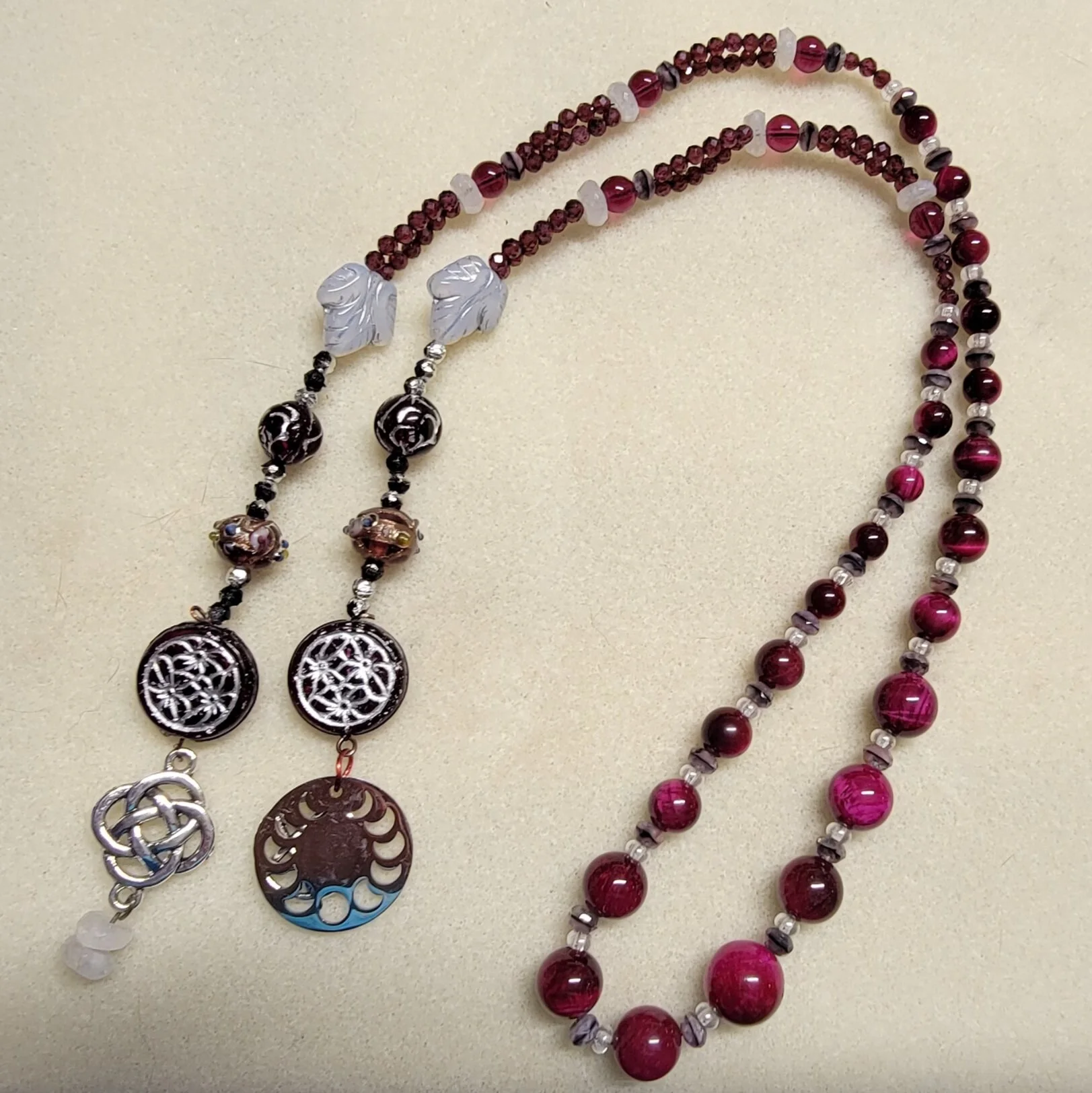 Handmade beaded necklaces with red, pink, and purple beads, Celtic knot and moon phase pendants, filigree charms, and leaf accents.