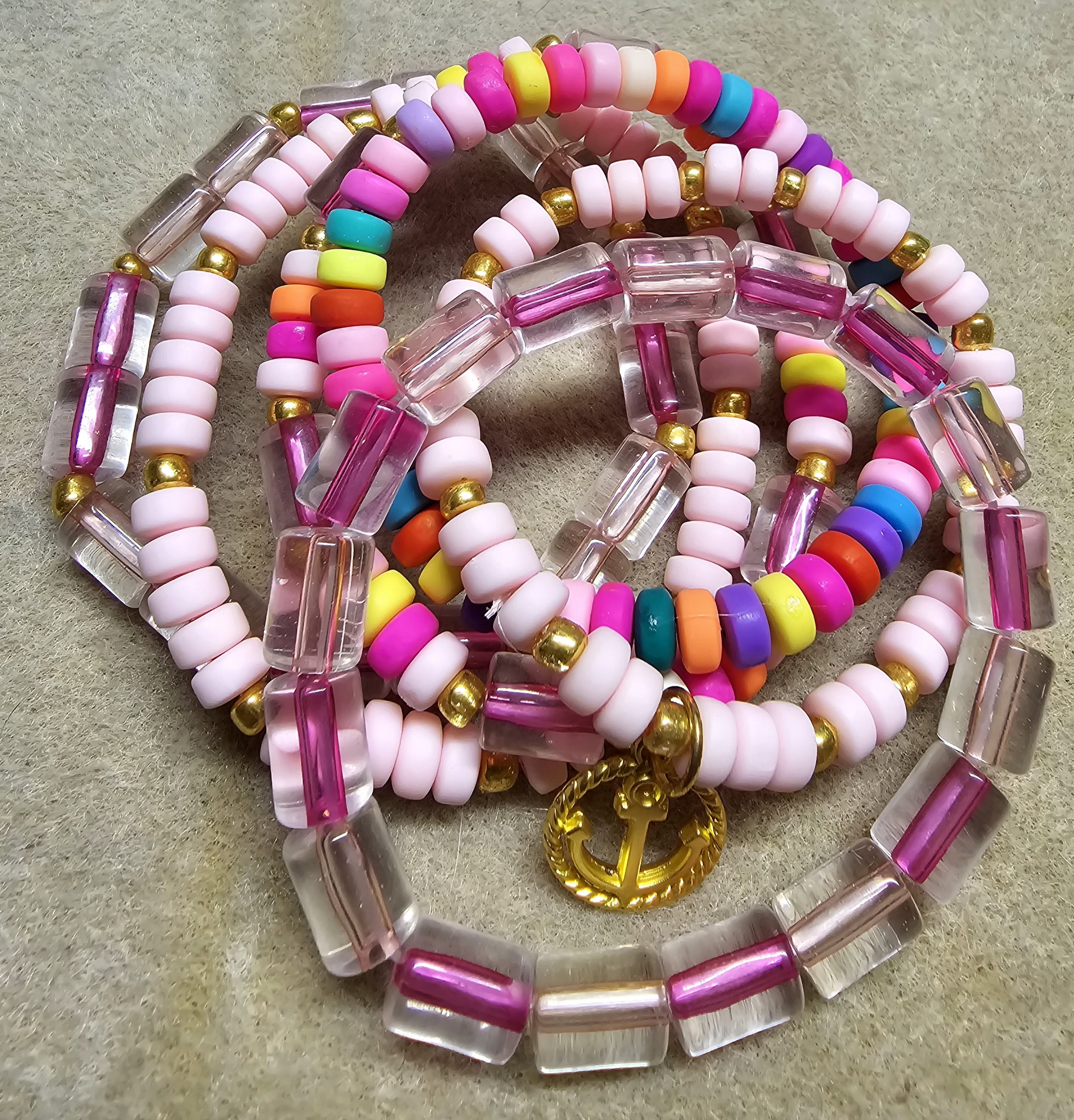 Colorful handmade stackable bracelets with pastel beads, gold spacers, and anchor charm on beige cloth