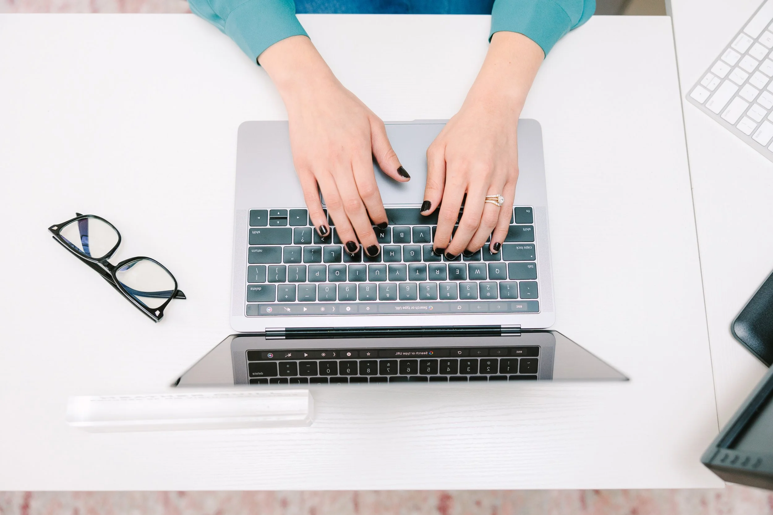Person typing on a laptop keyboard with glasses and other electronic devices on a white desk.