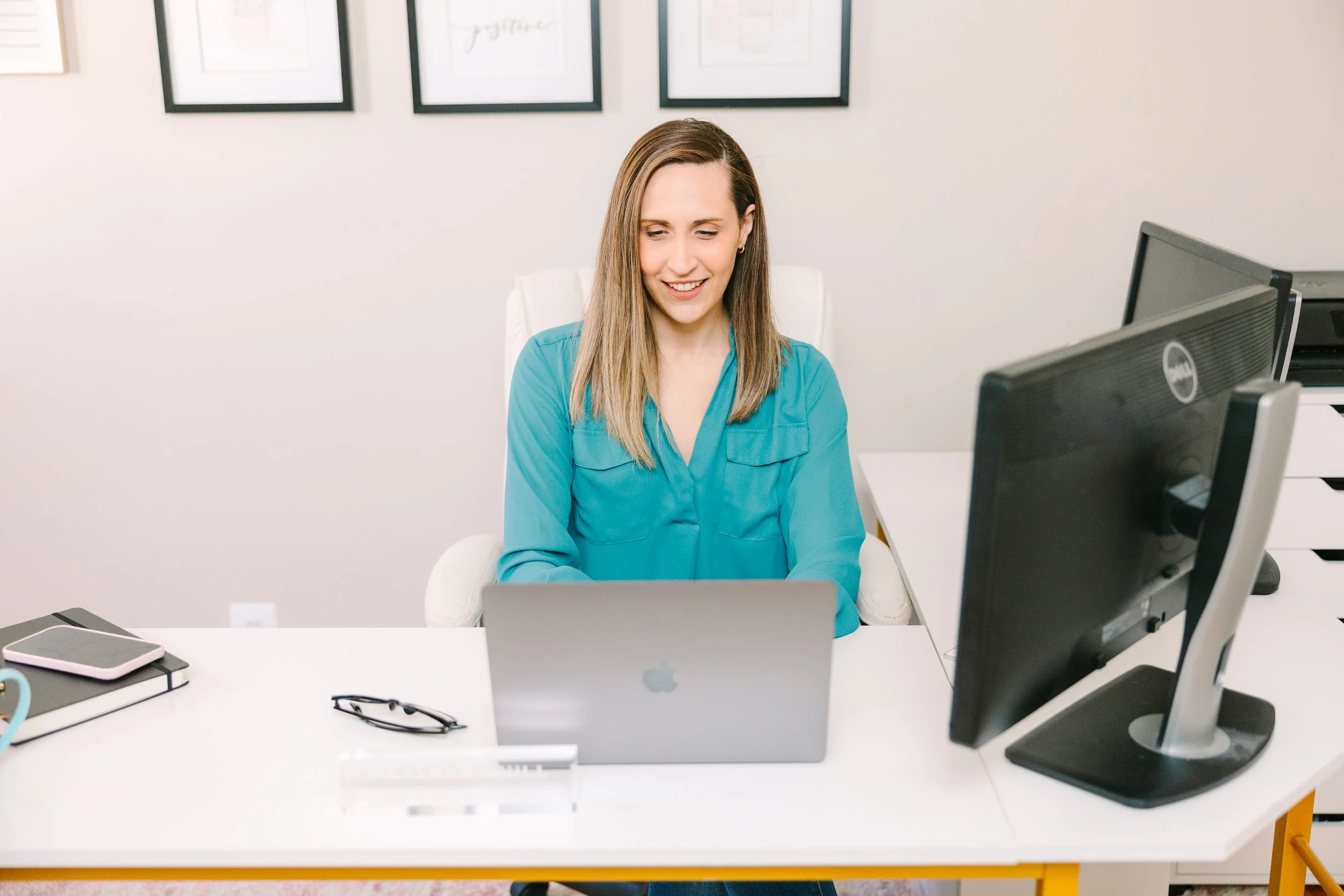 A woman with blonde hair working on a silver MacBook at an office desk, with a computer monitor and other office supplies around her.