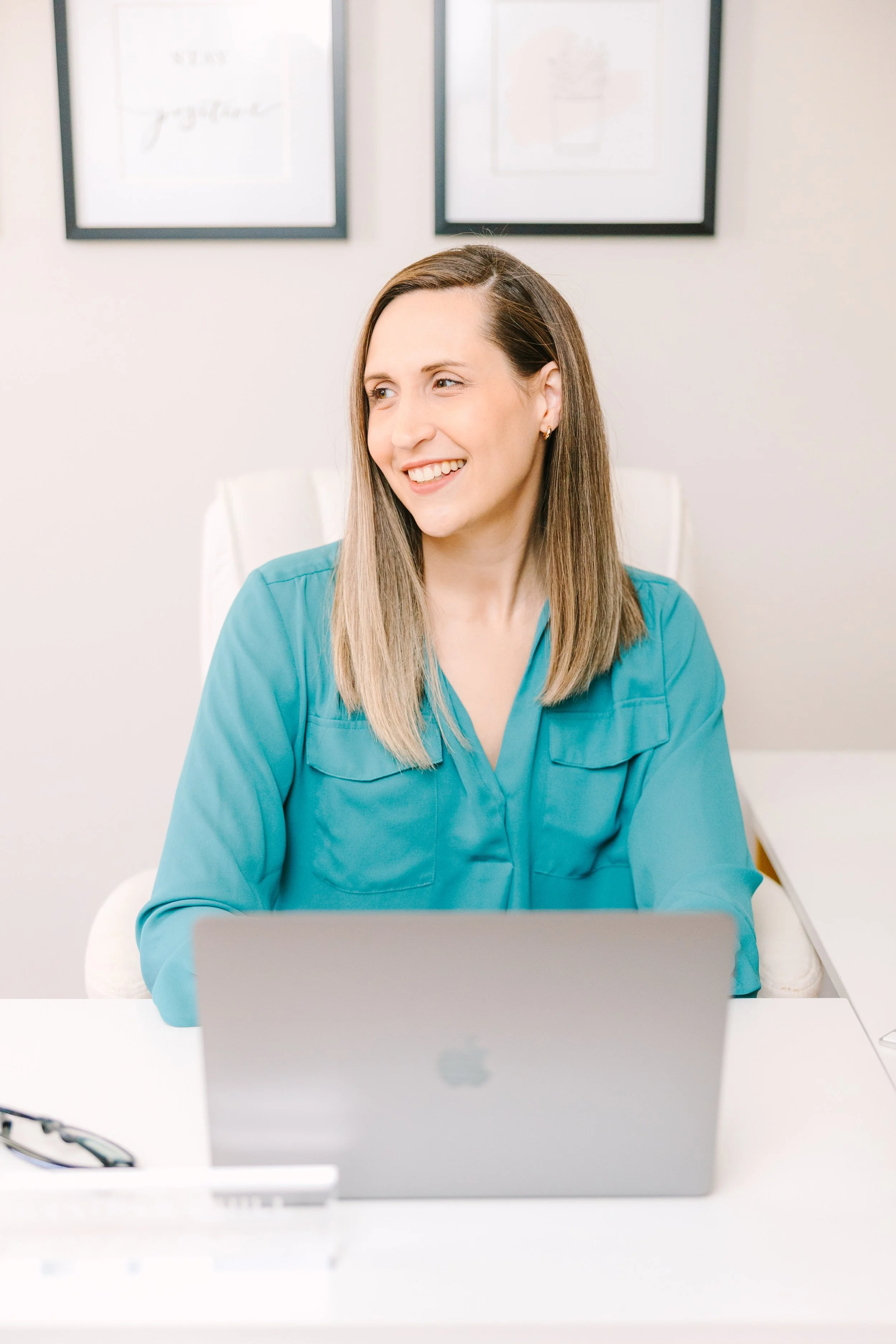 A woman with brown hair wearing a turquoise blouse sitting at a white desk with a silver MacBook.