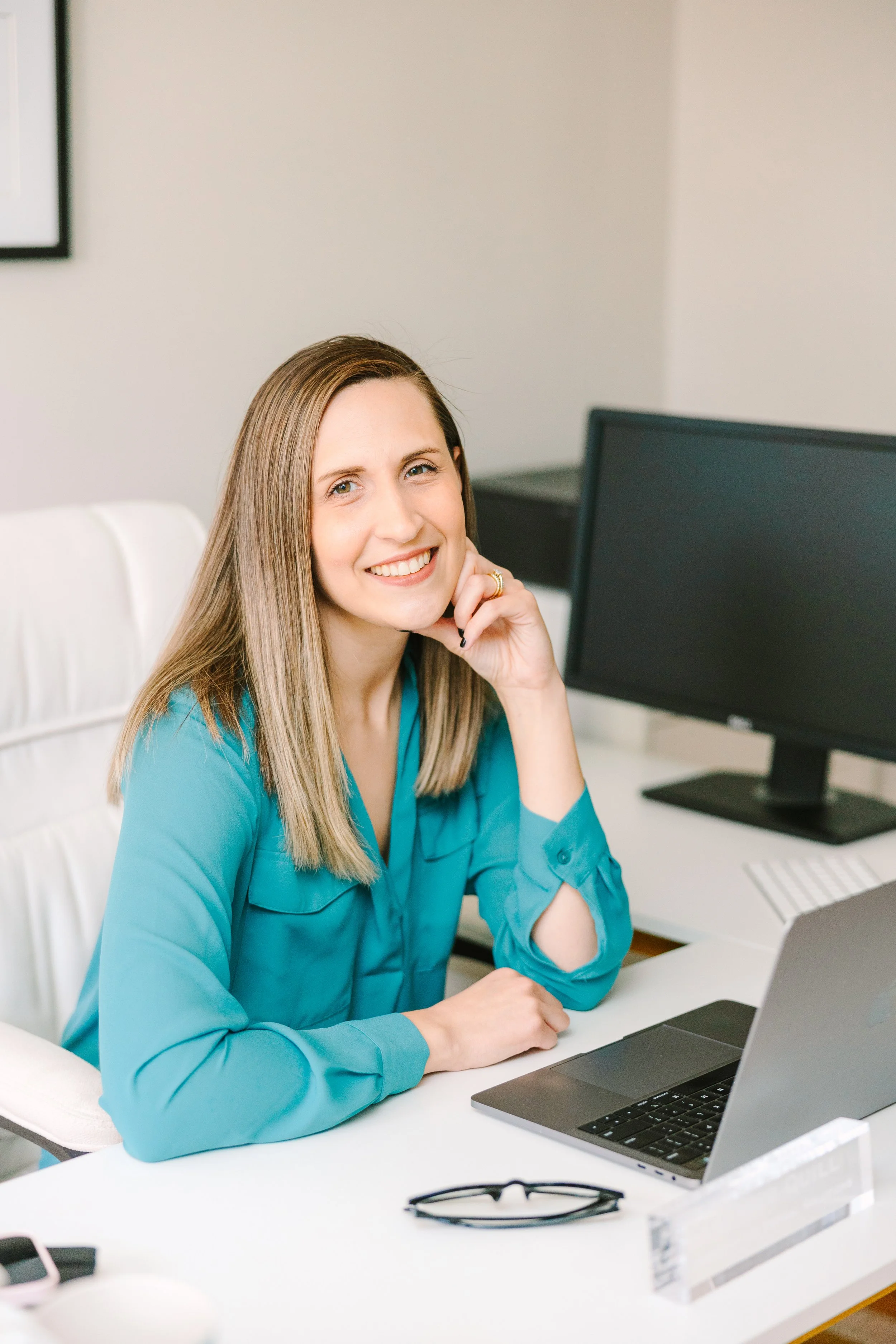 A woman with long blonde hair and a bright smile sitting at a desk in front of a laptop, wearing a teal blouse, with glasses and a computer monitor in the background.