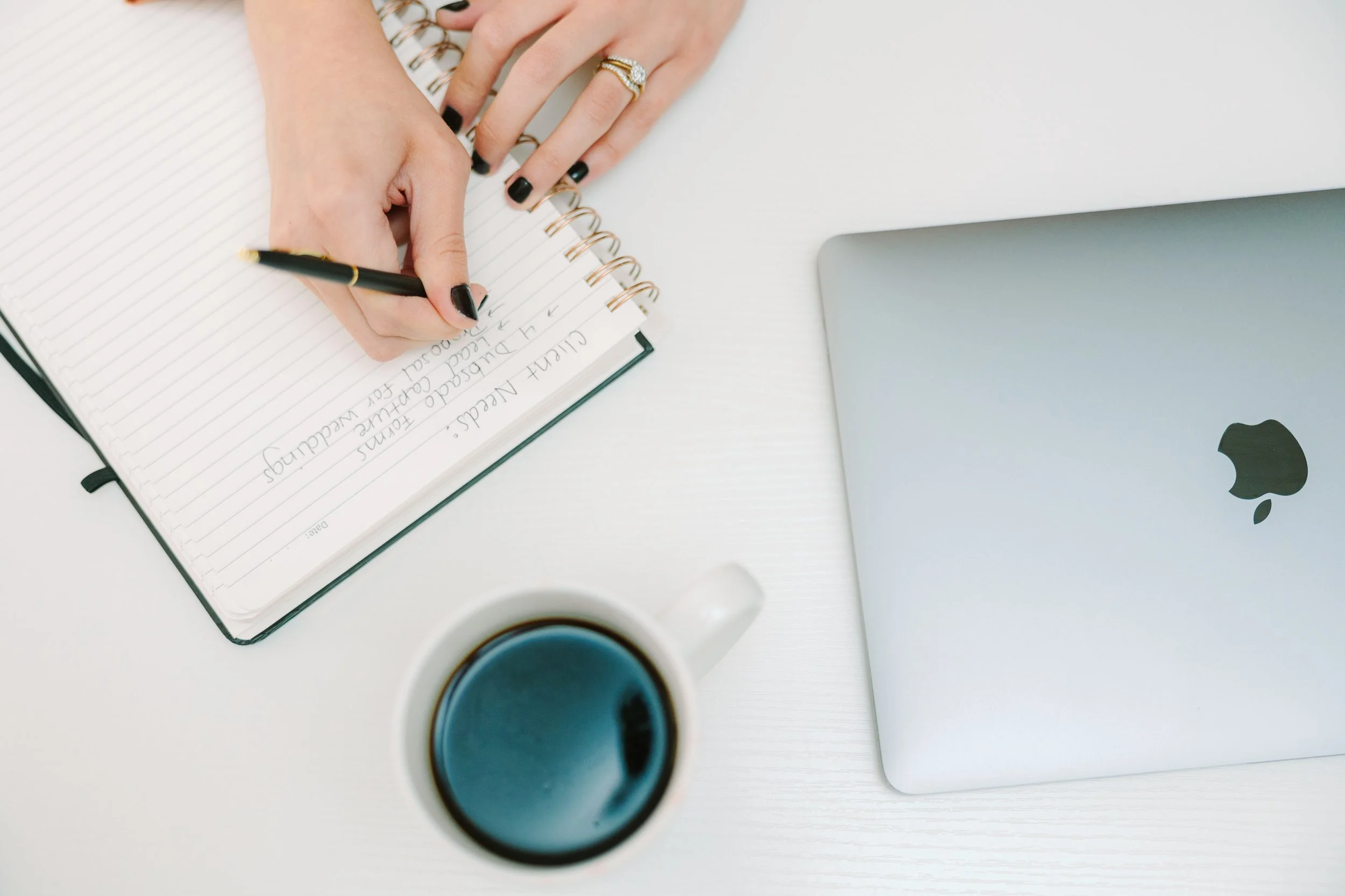 A person writes in a spiral notebook with a black pen on a white desk. Next to the notebook is a silver MacBook laptop with the Apple logo. A white cup of black coffee is also on the desk.