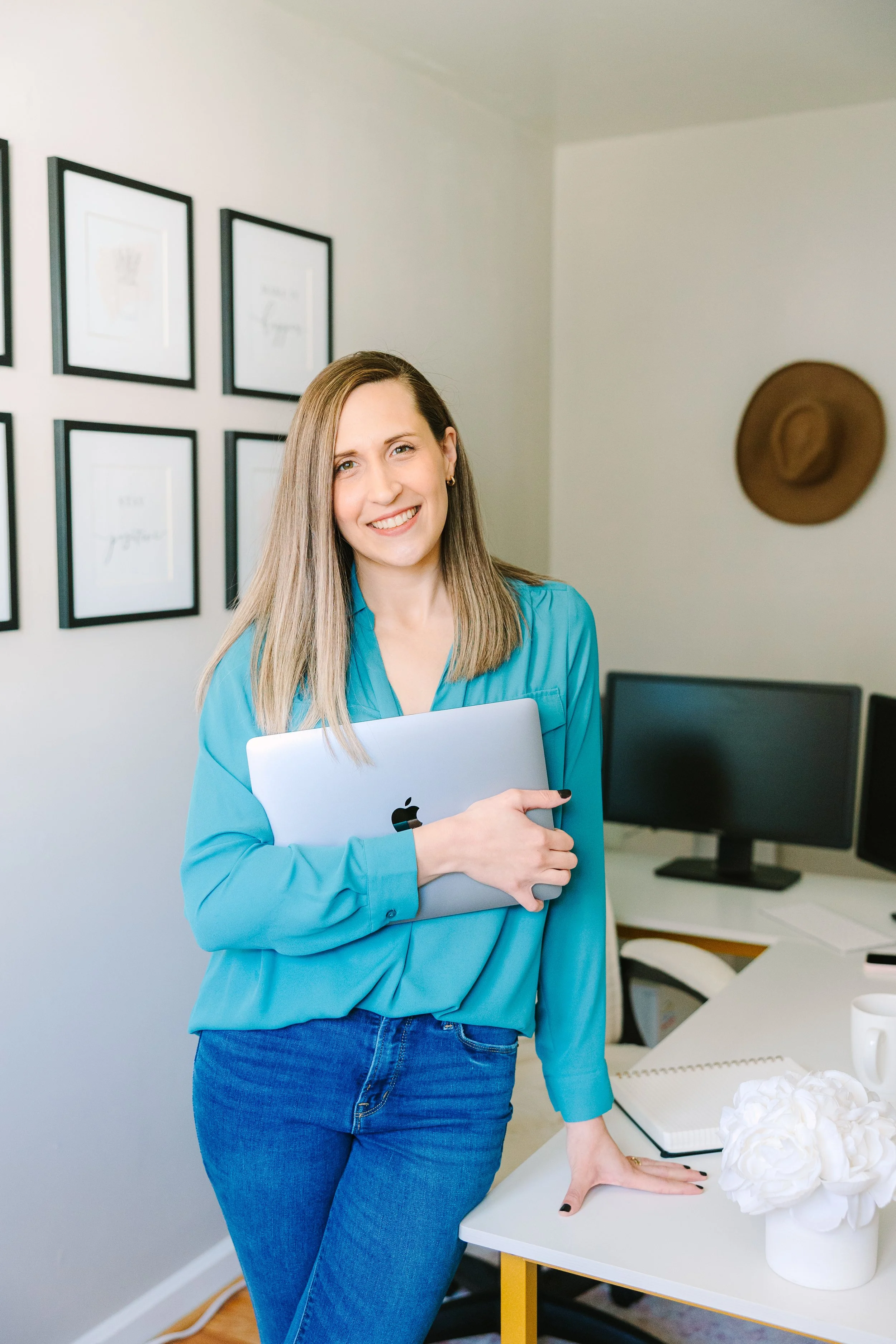 A woman in a blue blouse and jeans holding a silver MacBook, standing in a modern home office with framed art on the wall, a desk with multiple monitors, a notebook, a mug, and a bouquet of white flowers.