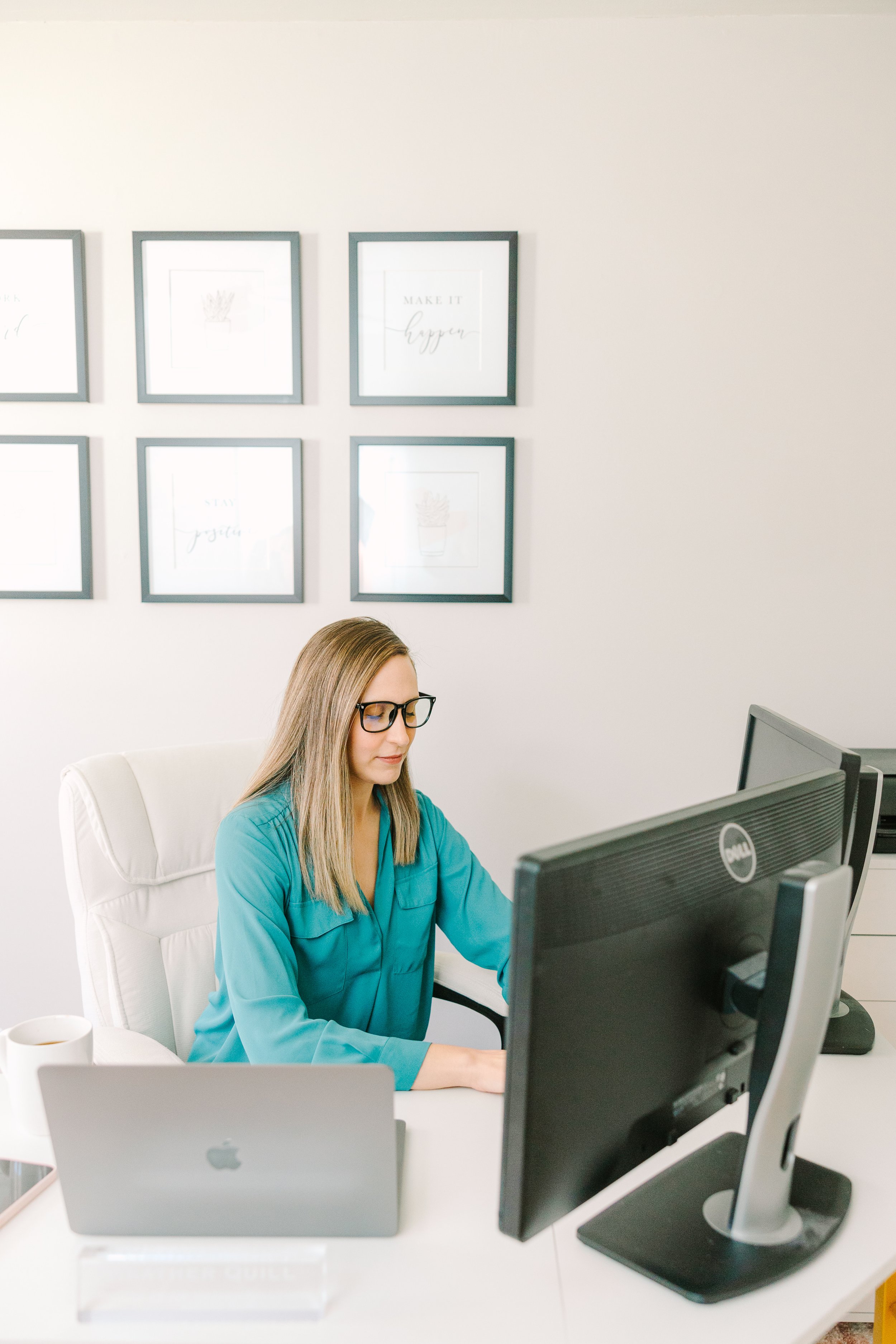 A woman with glasses and long blonde hair working at a desk in an office, with framed pictures on the white wall behind her.