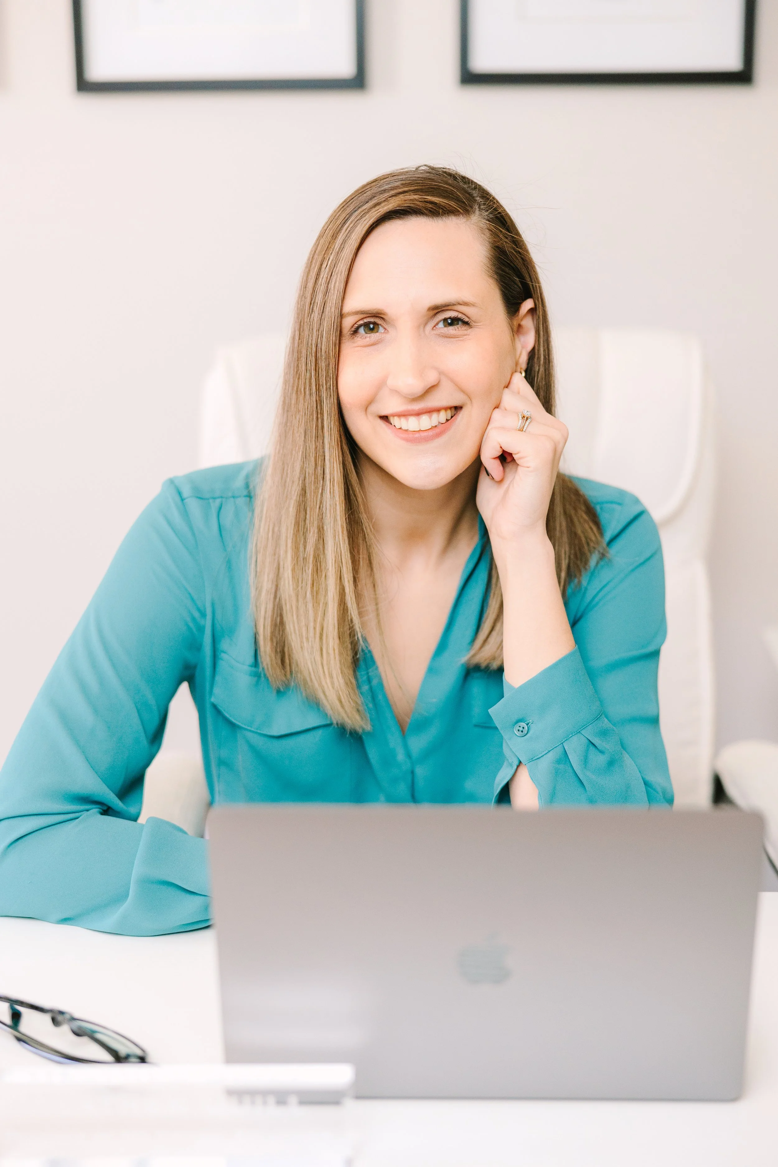 Smile woman in a blue shirt sitting at a white desk with a silver laptop in front of her.