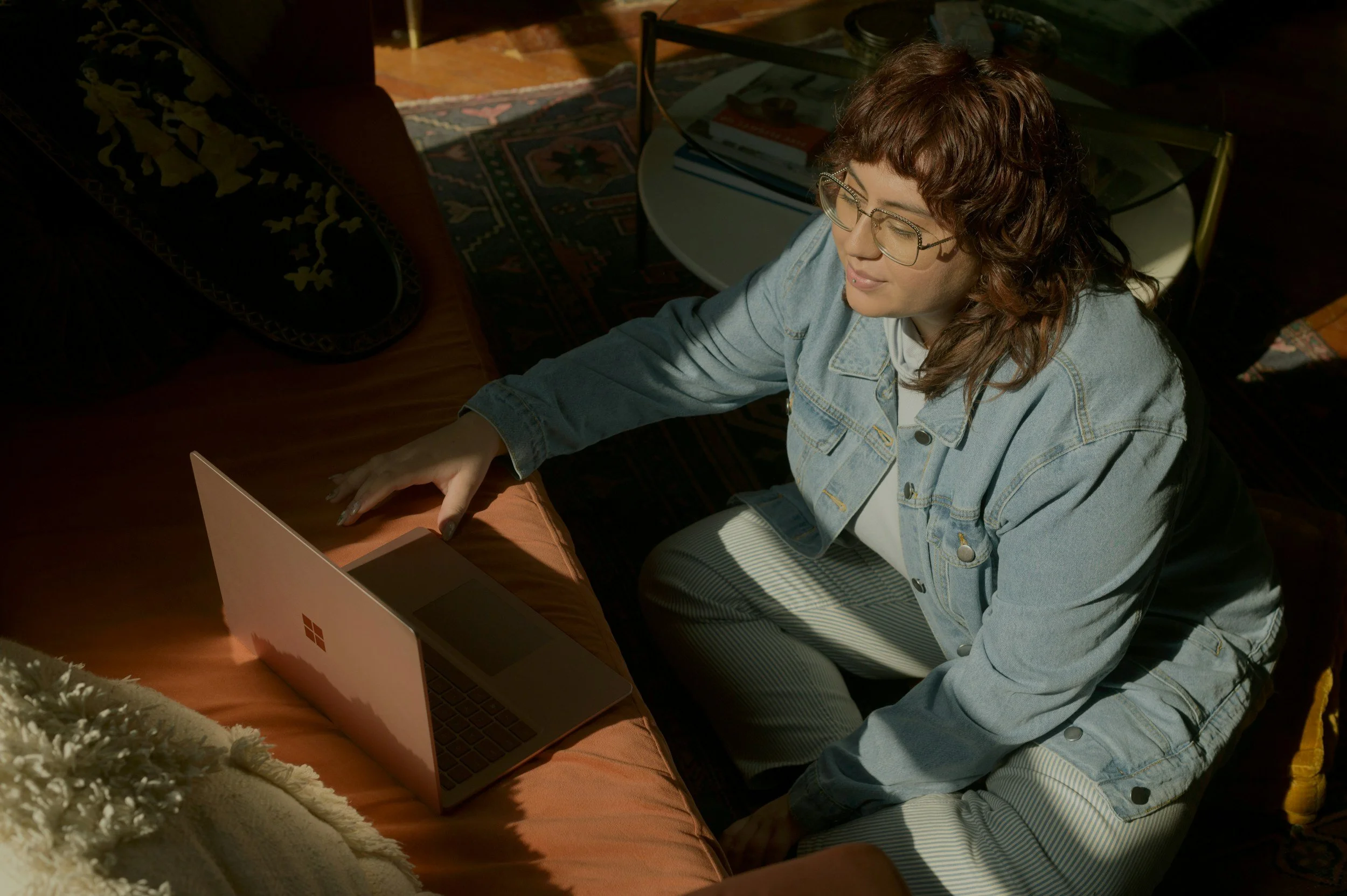 A woman with curly brown hair, wearing glasses, a light blue denim jacket, and striped pants, sitting on the floor and working on a silver laptop with a red window logo, in a dimly lit room with a patterned rug, a round glass table with books, and a sofa and pillow visible in the background.