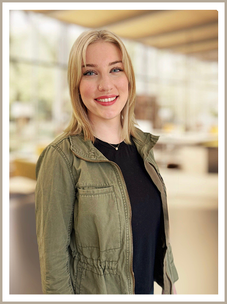 Young woman smiling indoors in a casual outfit, wearing a green jacket over a black shirt.