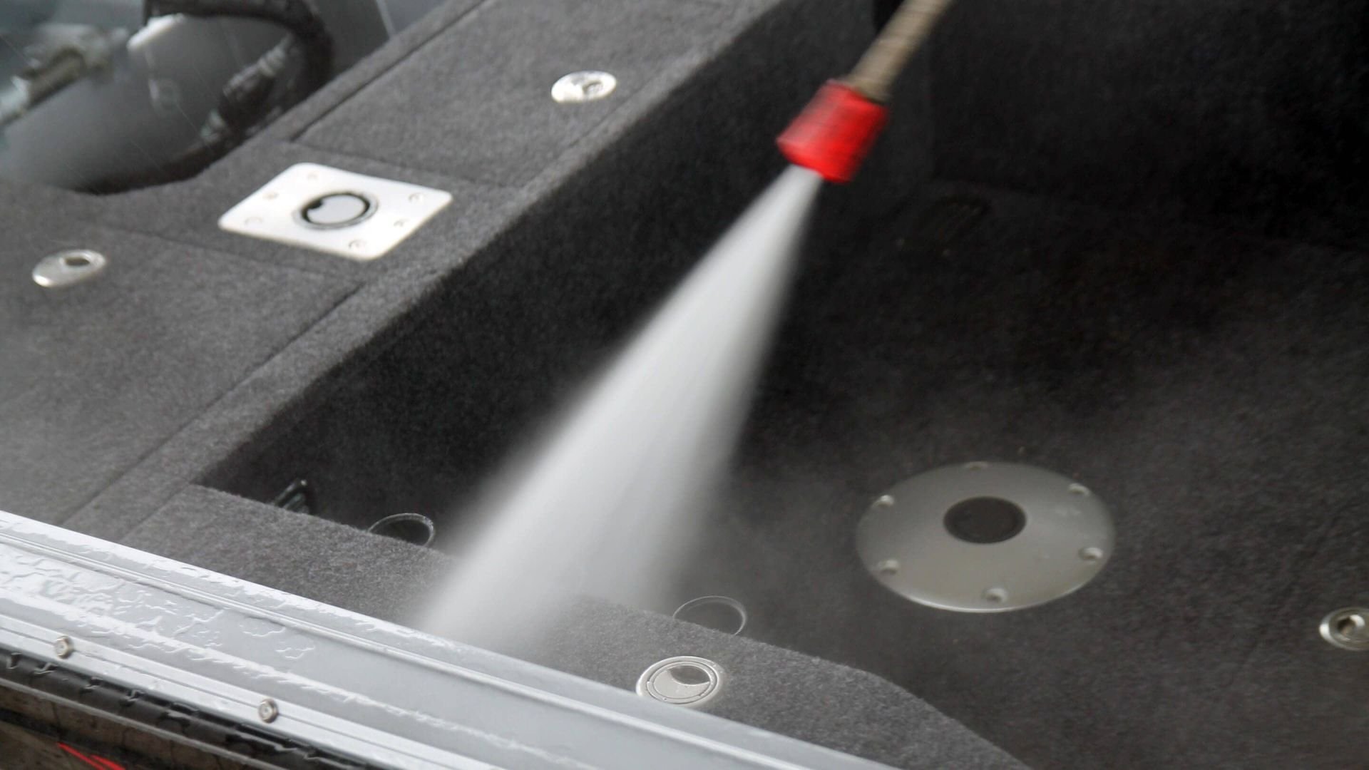 A spray of water being used to clean or rinse a black carpeted surface with metal mounting points.