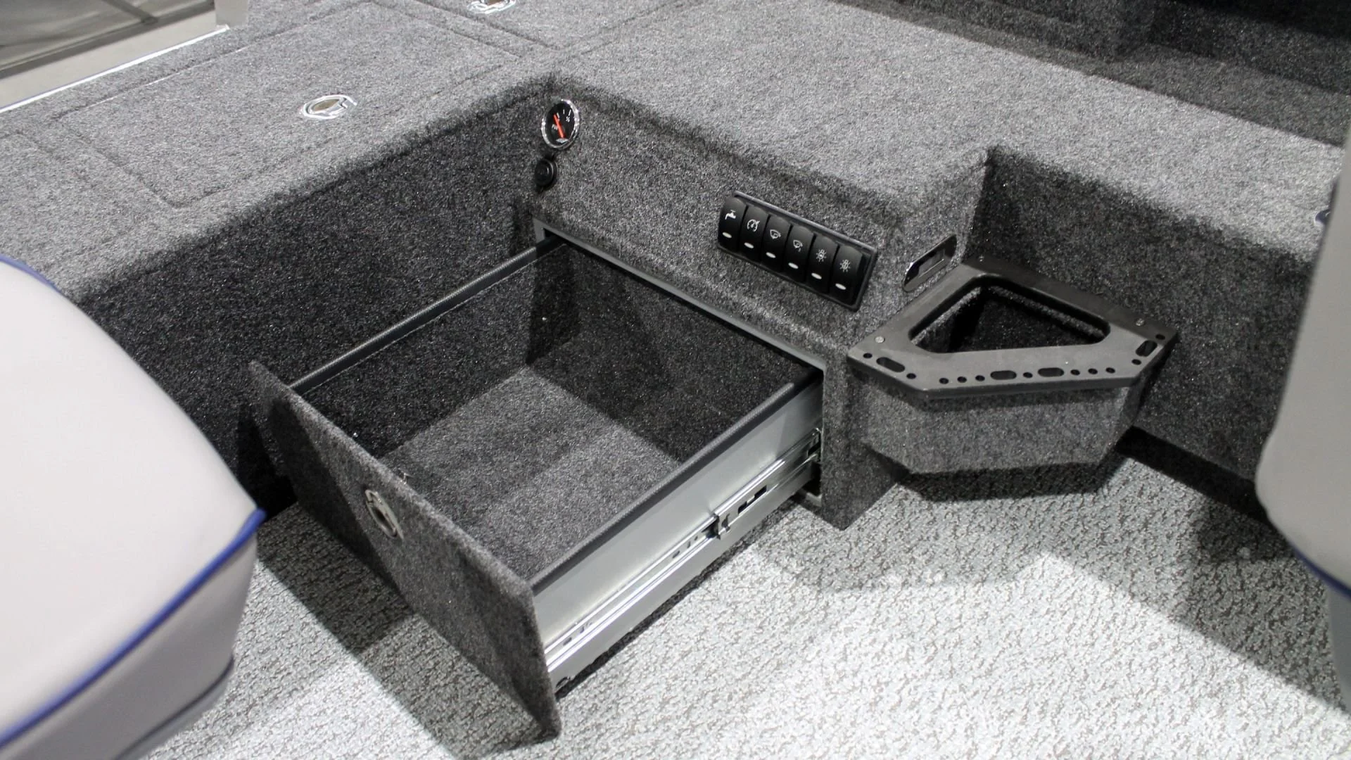 Interior of a vehicle with a storage compartment built into the floor, surrounded by gray carpet. There is a control panel with switches, a gauge, and a bracket for holding items.
