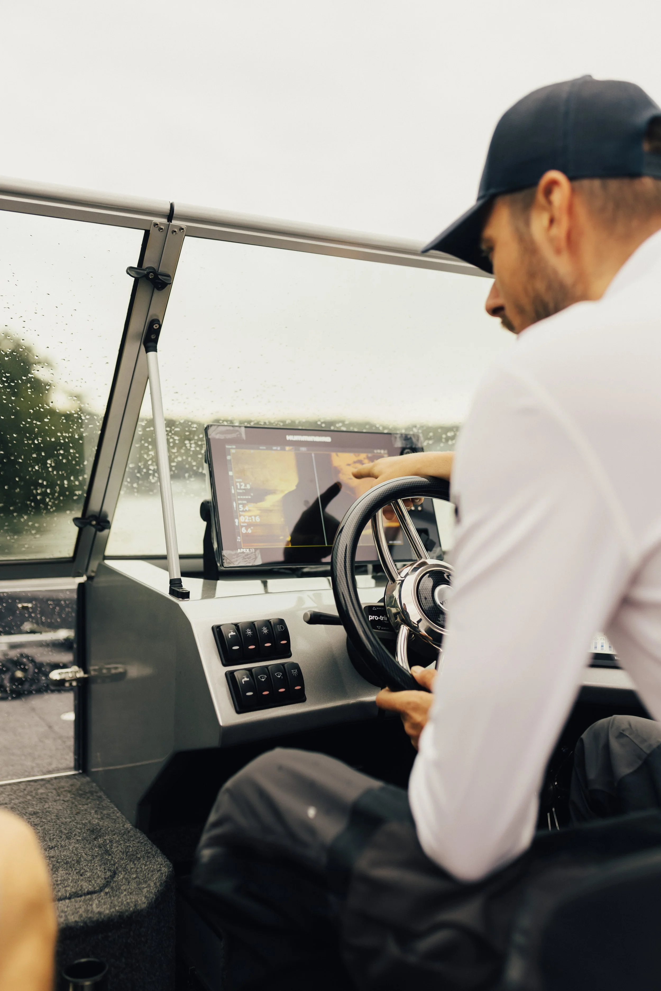 A man wearing a black cap and white shirt sitting inside a boat, steering with his right hand while pointing at a GPS screen with his left hand on a rainy day.