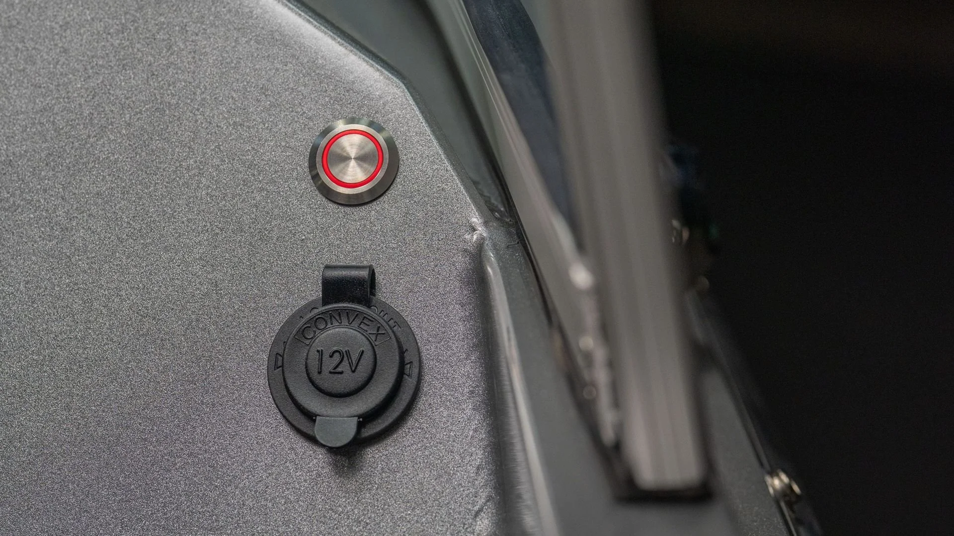 Close-up of a 12V power outlet and a red button on a textured metallic surface.