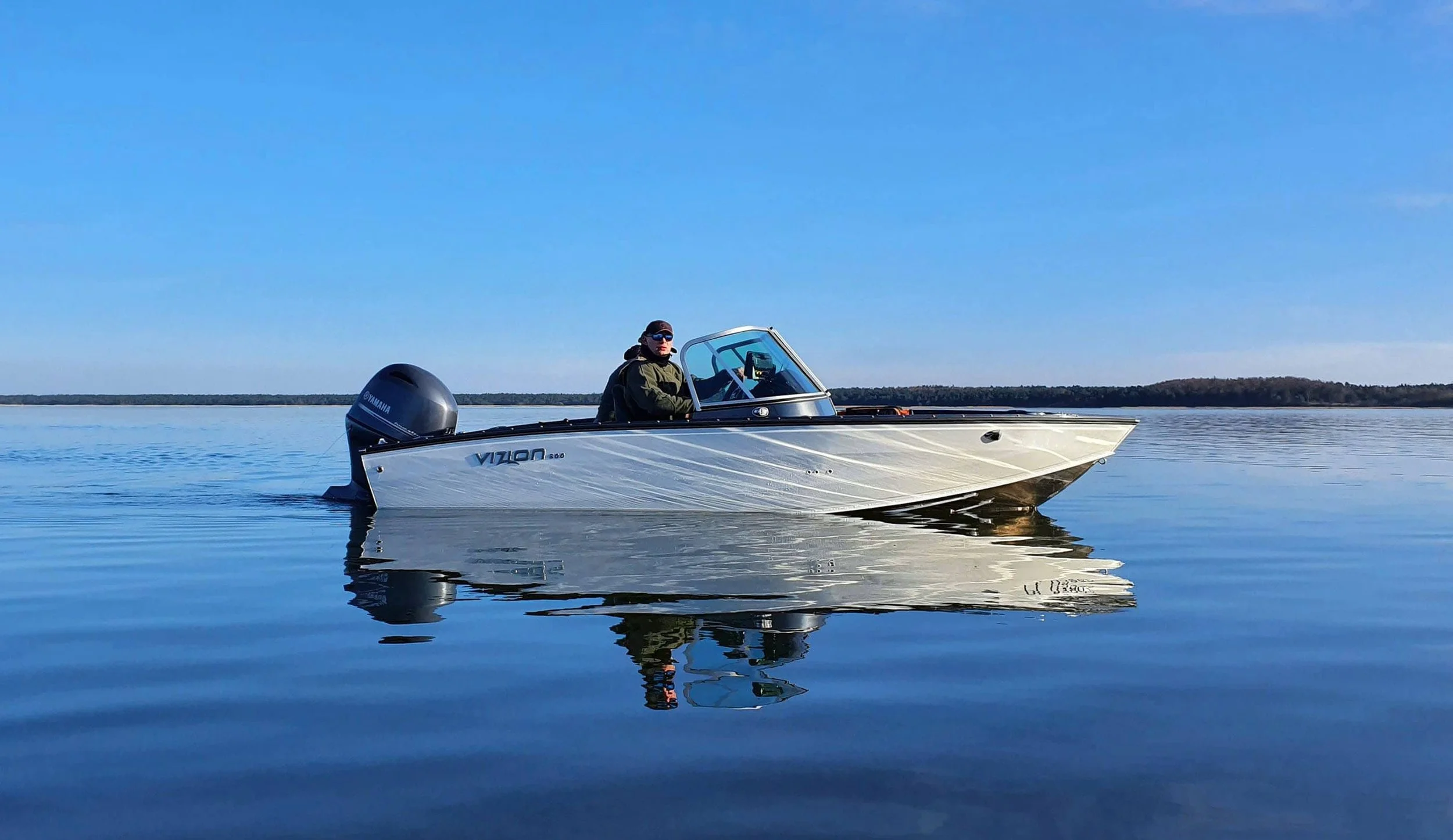 A person in sunglasses and a jacket driving a small motorboat on calm water under a clear blue sky.