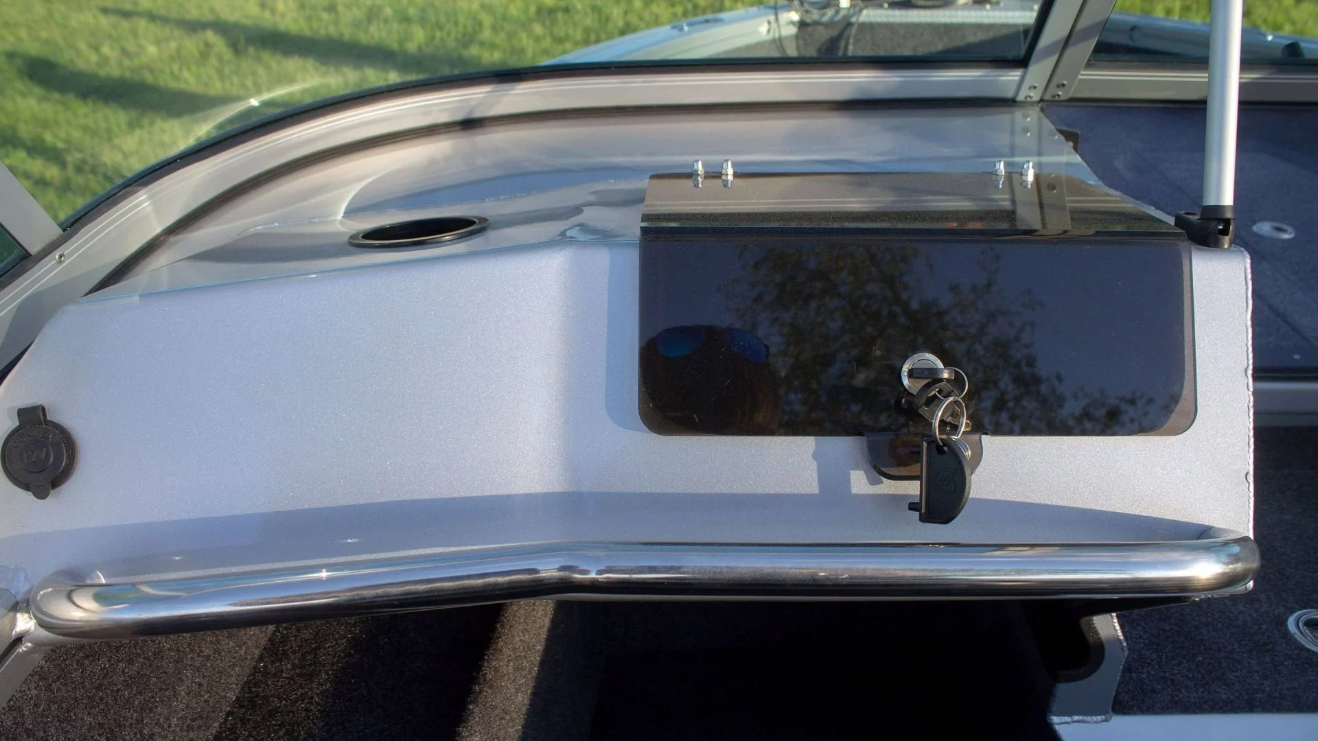 Close-up of a boat's dashboard showing a black rectangular panel with a lock and key, a mirror, and a circular cover on the side. The background includes a glimpse of water and greenery.
