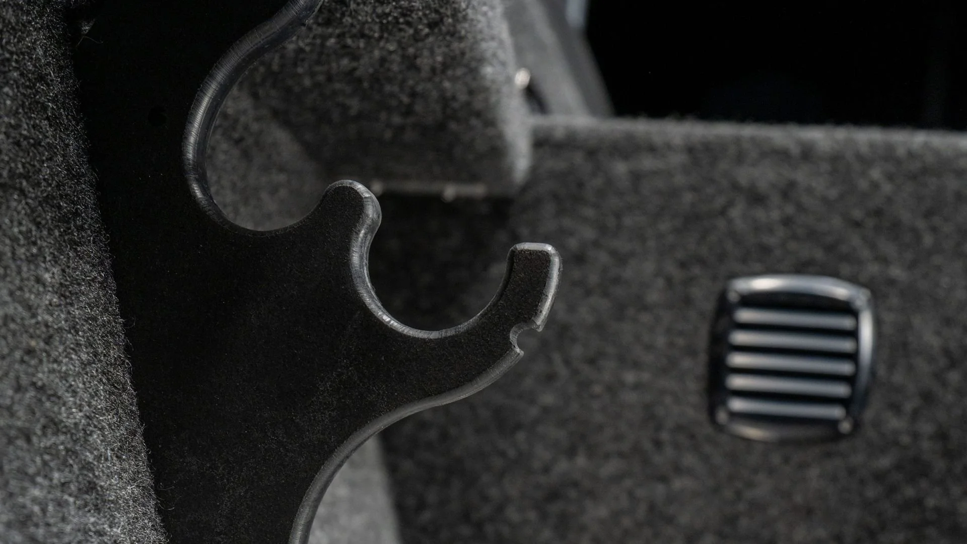 Close-up of a black hook-shaped object, a foot pedal, and carpeted surface inside a vehicle.