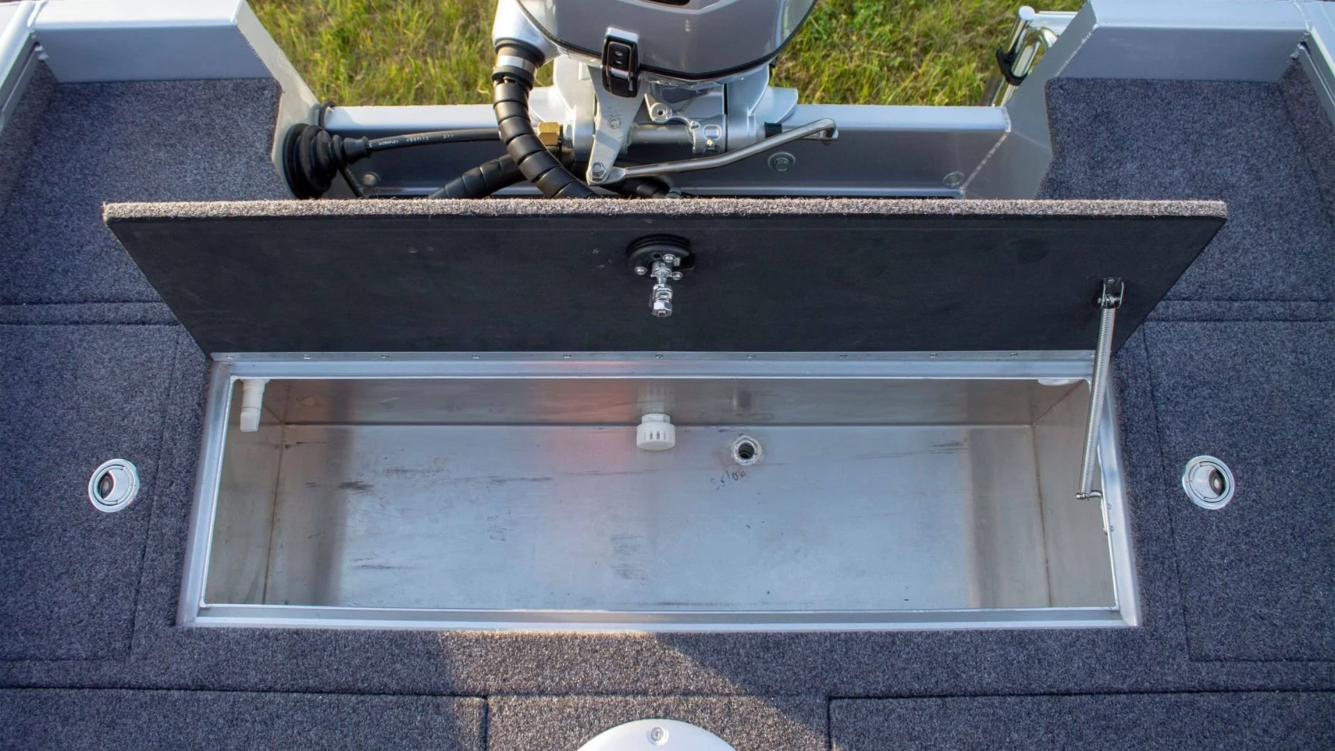 Open storage compartment on a boat with a cover, showing an empty metal interior with a drain hole and a white pipe fitting, surrounded by gray carpeting.
