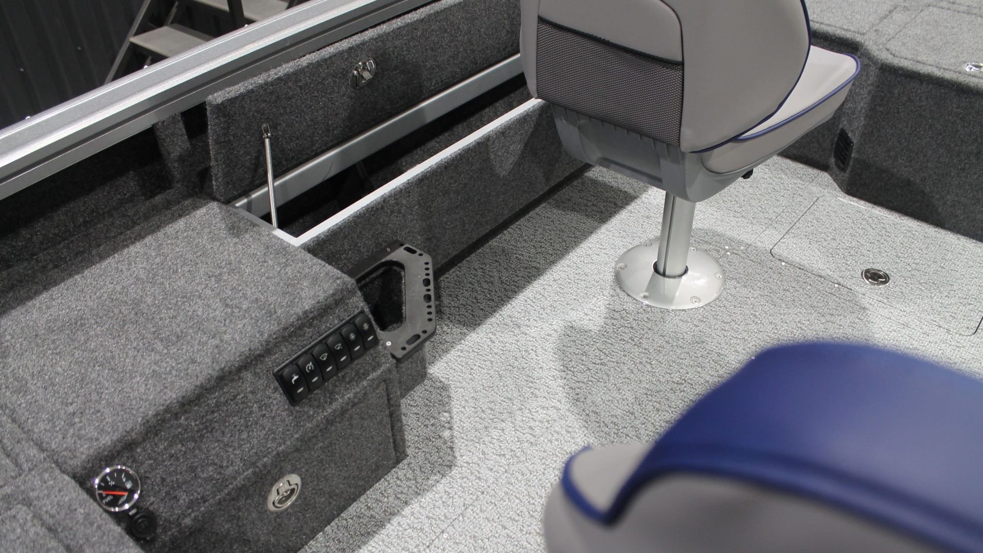 Interior of a boat showing a gray chair with a rounded backrest and base, a control panel with switches, and a storage compartment with a gauge. The floor is carpeted, and there are storage hatches and a railing.